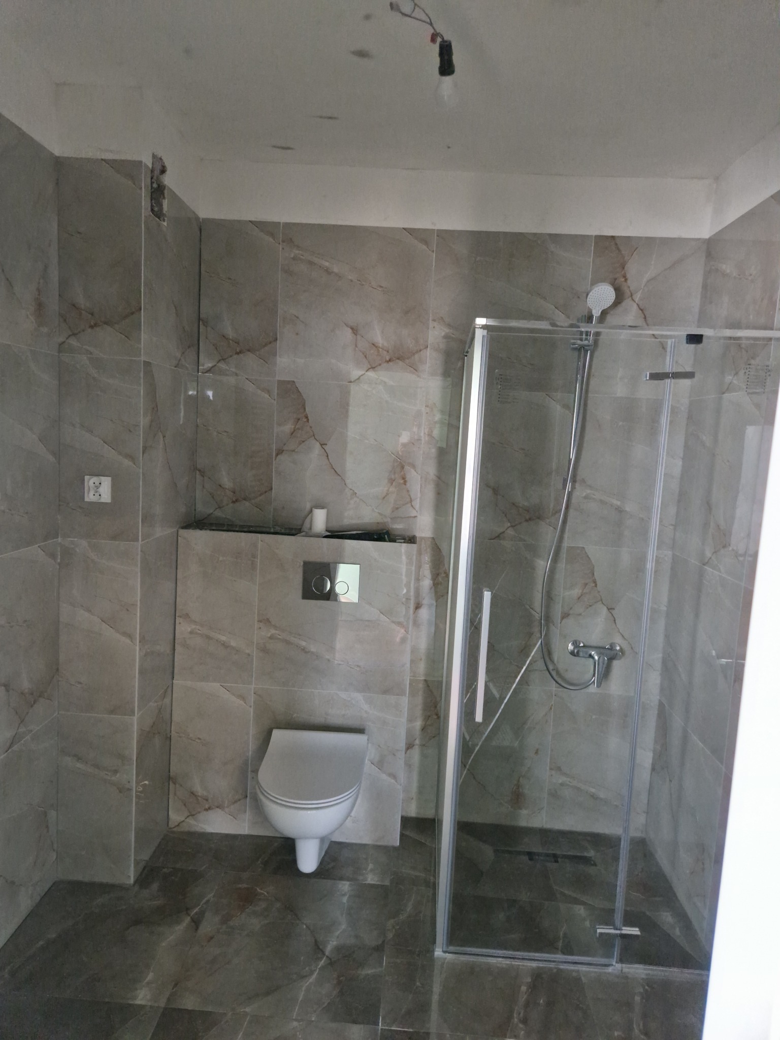 Wykończona łazienka z marmurowymi płytkami, szklaną kabiną prysznicową i podwieszaną toaletą, widoczne niedociągnięcia w górnym rogu ściany, surowa żarówka zwisająca z sufitu.