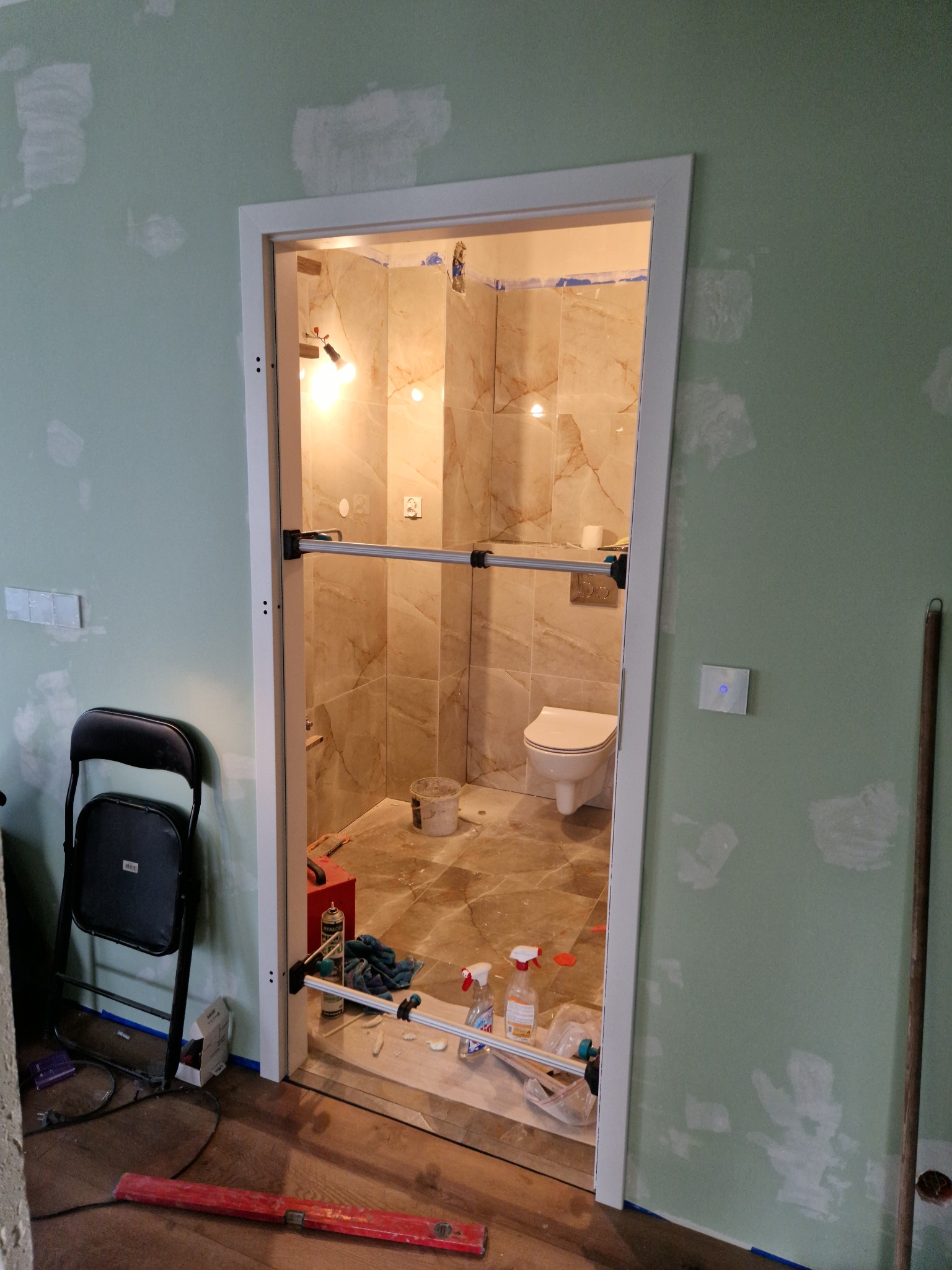 Łazienka w trakcie prac wykończeniowych: widoczne płytki imitujące marmur, biała toaleta, narzędzia i materiały budowlane, ściany przygotowane do malowania, zamontowane drzwi z zabezpieczeniem...