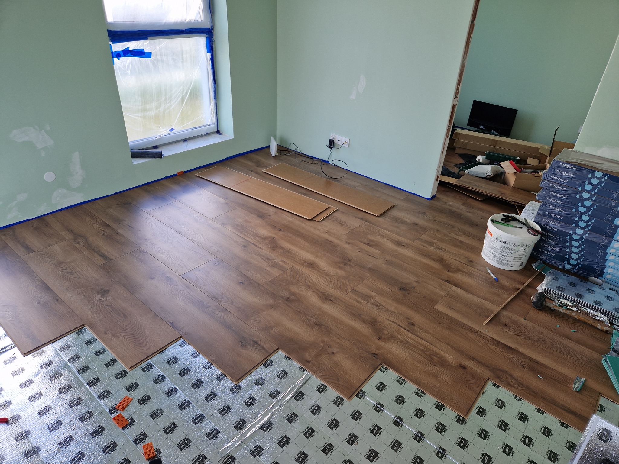 Proces układania paneli podłogowych w pokoju z jasnymi ścianami i oknem zabezpieczonym folią malarską; widoczne panele, podkład izolacyjny z nadrukiem 'CUTTING LINES' oraz opakowania paneli Megaloc.