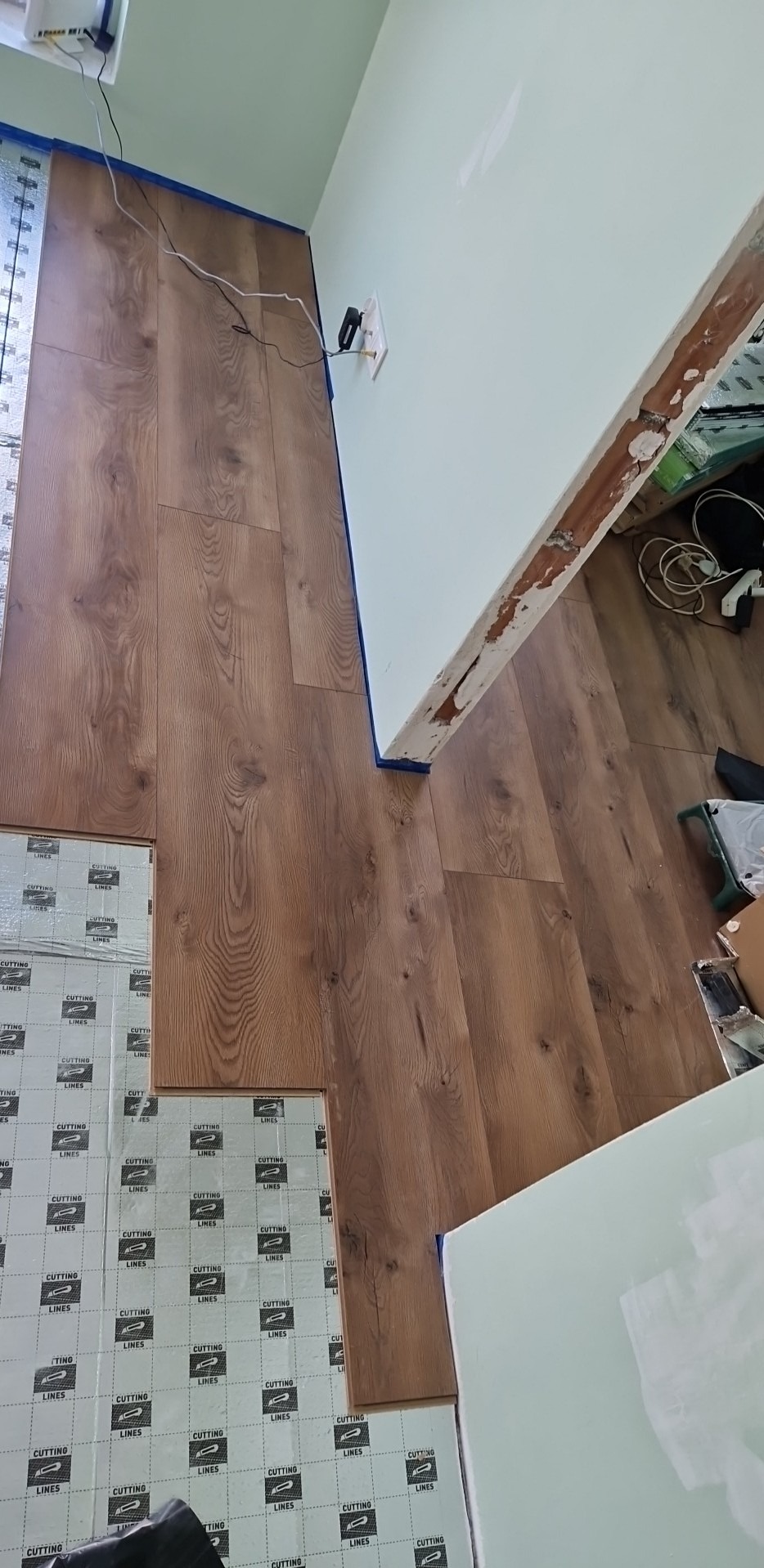 Panele podłogowe o wzorze drewna w trakcie układania, widoczne podkłady z nadrukowanymi liniami cięcia, niebieska taśma malarska przy ścianie, fragment niezabezpieczonej futryny drzwi.