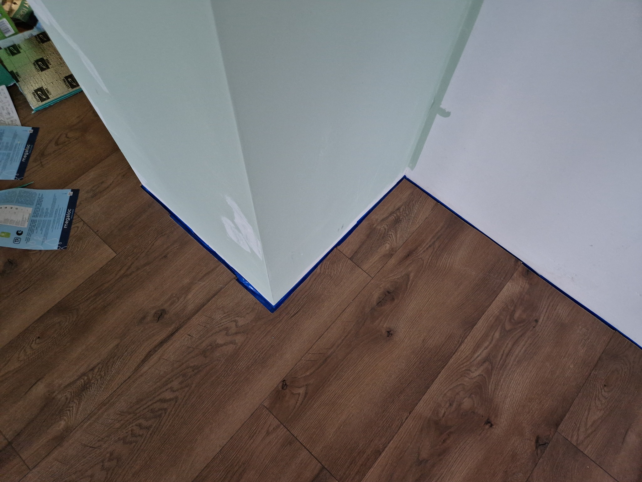 Ukończone układanie paneli podłogowych w jodełkę, zabezpieczone niebieską taśmą malarską przy ścianach pomalowanych na jasnozielony kolor, widoczne paczki paneli w tle.