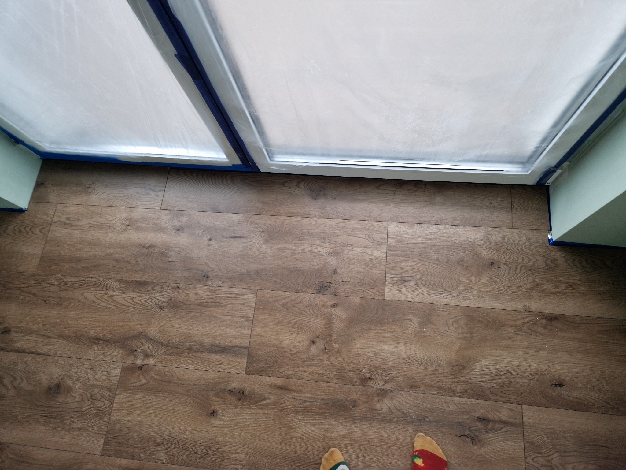 Podłoga z paneli w odcieniach brązu, zabezpieczona folią i niebieską taśmą malarską przy oknie, widoczne fragmenty skarpetek.