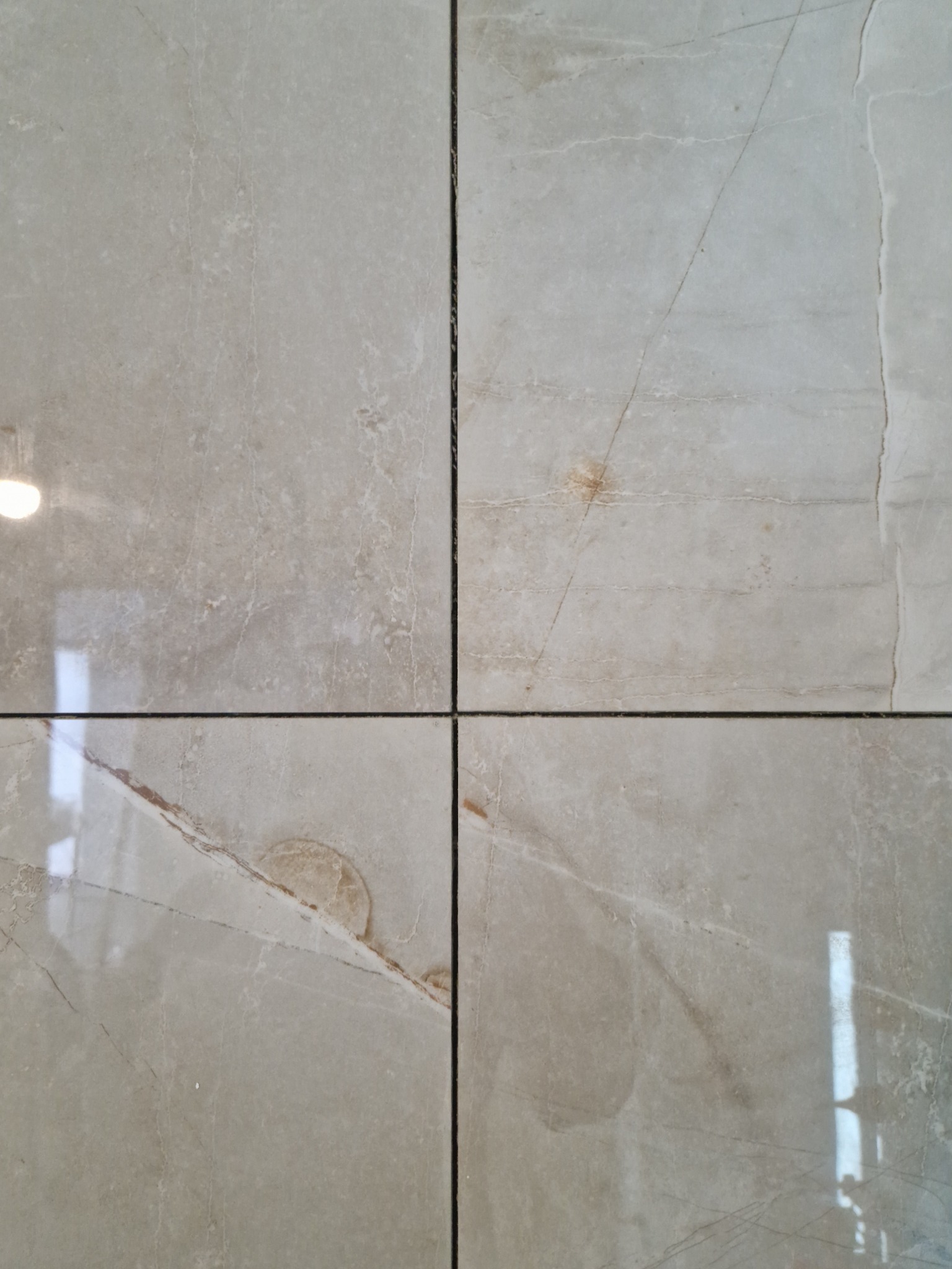 Cztery beżowe płytki podłogowe z marmurkowym wzorem, ułożone z wąskimi, ciemnymi fugami, odbijające światło w górnej lewej i dolnej prawej płytce.
