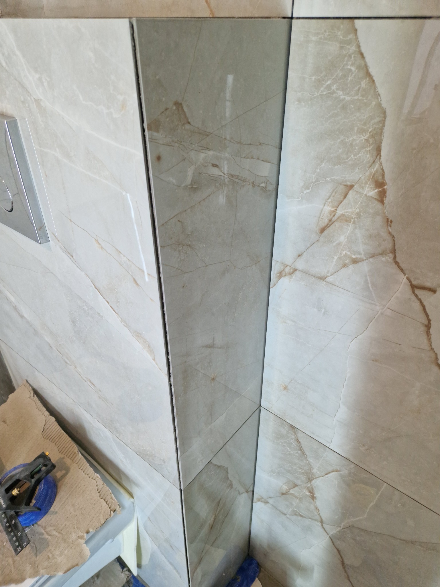 Układanie płytek imitujących marmur w Toruniu, widoczny fragment instalacji podtynkowej WC, narzędzie glazurnicze na kartonie.