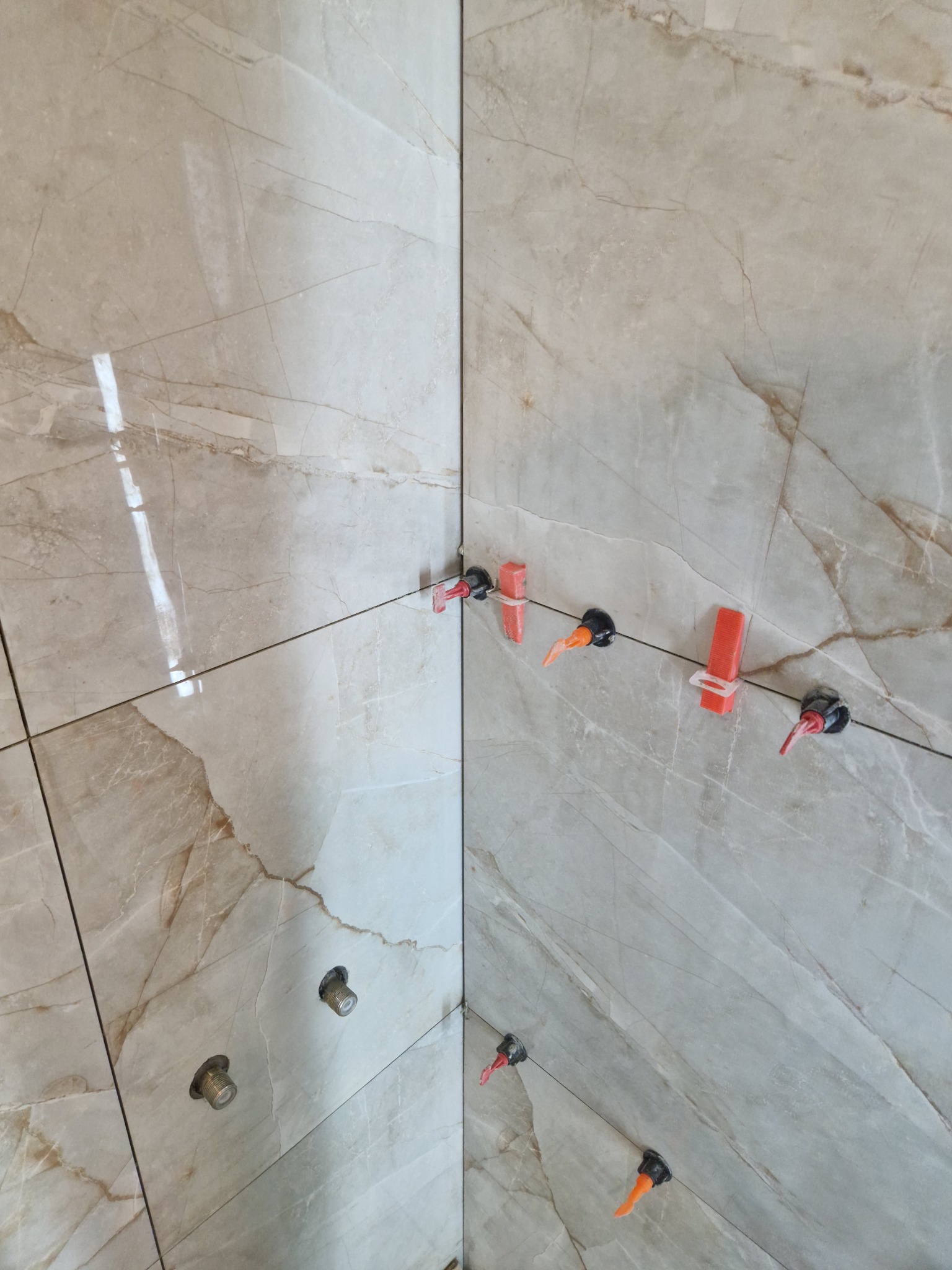 Układanie beżowych płytek imitujących marmur na ścianie, widoczne plastikowe klipsy i kliny do poziomowania, otwory pod baterie.