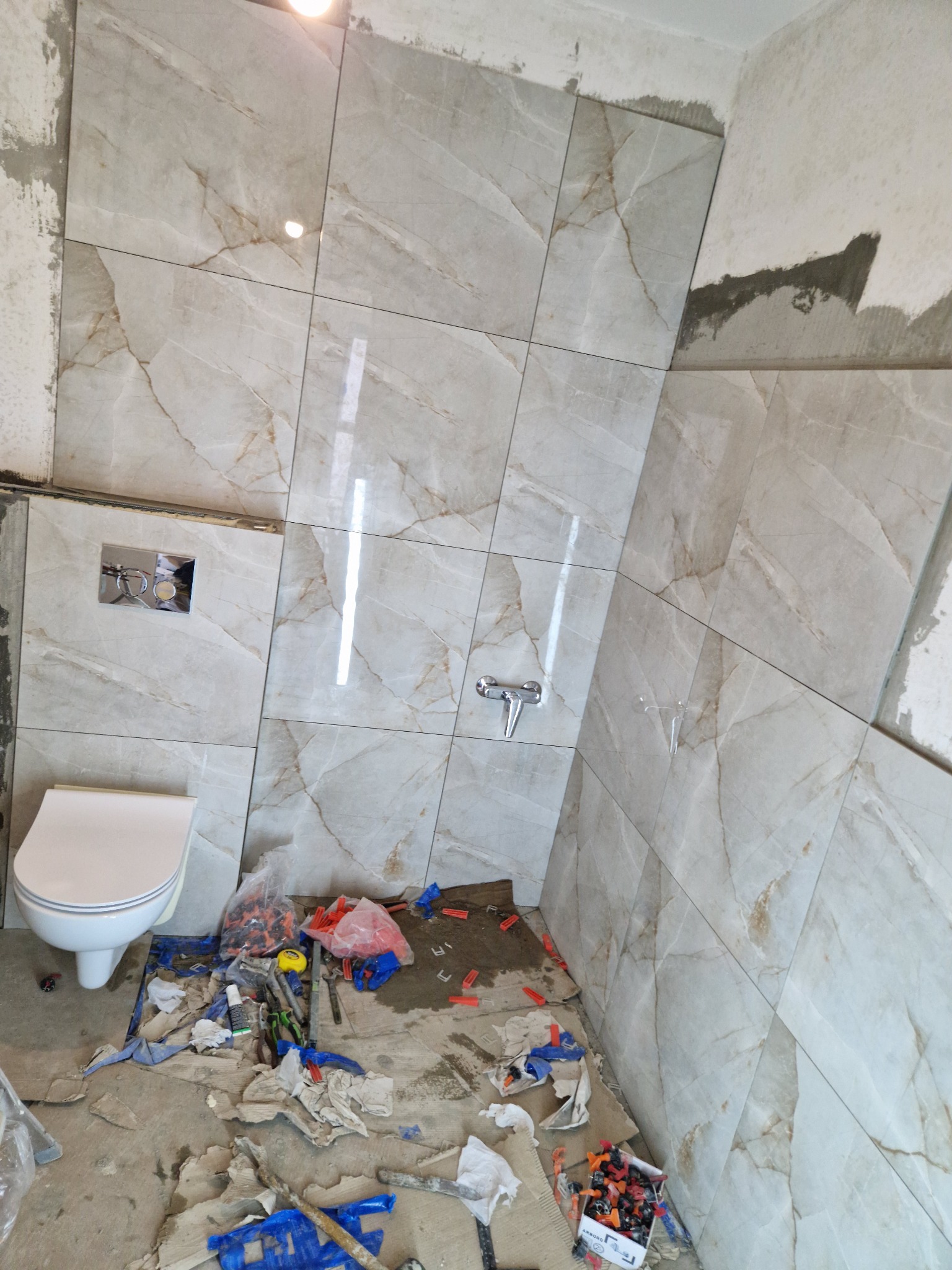 Wnętrze łazienki w trakcie prac: ułożone marmurowe płytki ścienne, zamontowana armatura, widoczne narzędzia i materiały glazurnicze na podłodze, częściowo zamontowana toaleta.