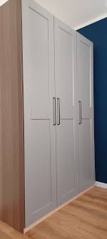 Szara, trzydrzwiowa szafa z ciemnymi uchwytami na tle niebieskiej ściany i drewnianej podłogi. Widoczny fragment brązowej bocznej ścianki szafy.