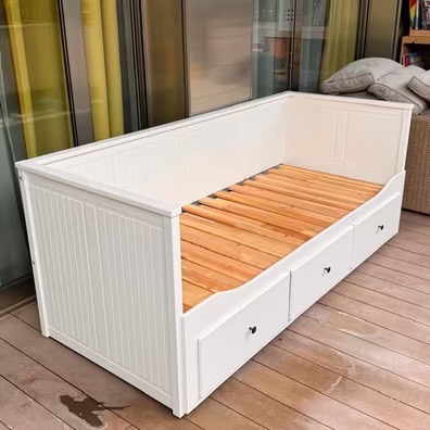 Biała, drewniana sofa z wysuwanymi szufladami i drewnianym stelażem, ustawiona na drewnianej podłodze. Widoczne elementy dekoracyjne w tle.