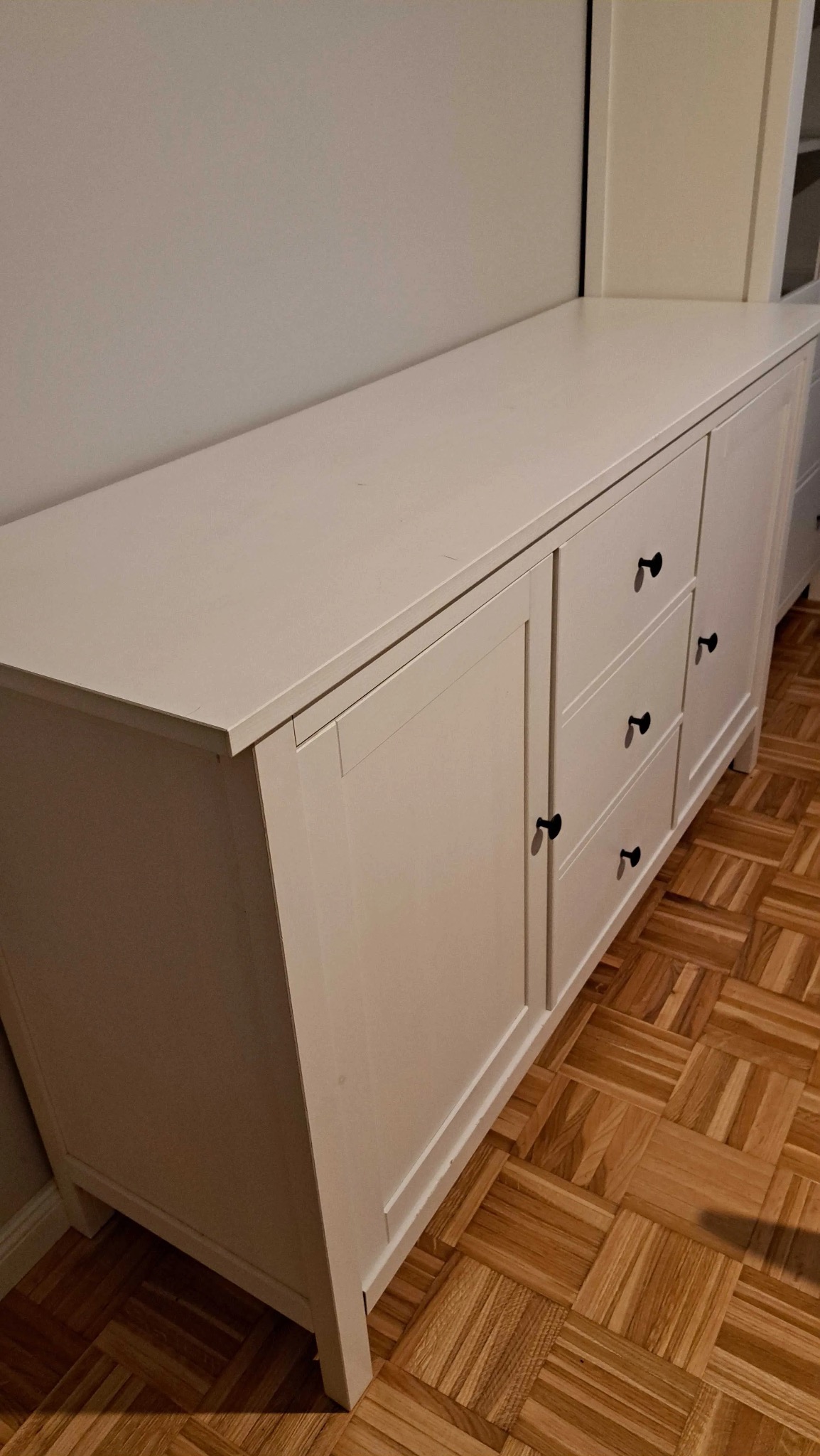 Biała komoda z trzema szufladami i dwojgiem drzwi, z czarnymi uchwytami, stoi na parkiecie w jodełkę. Widok z boku, oświetlenie naturalne.