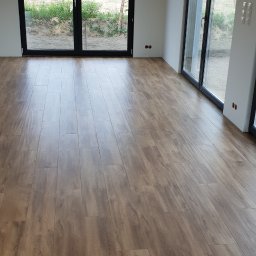 KONKRET Zbigniew Stańczak - Ułożona podłoga z paneli imitujących drewno w jasnym, pustym pokoju z dużymi, czarnymi oknami balkonowymi, widoczne kable wystające z sufitu i gniazdka w ścianach.