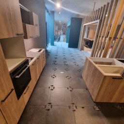 KONKRET Zbigniew Stańczak - Montaż szarej podłogi z płytek w kuchni, widoczne klipsy poziomujące, częściowo zamontowane szafki z drewnopodobnym wykończeniem, drabina w tle.