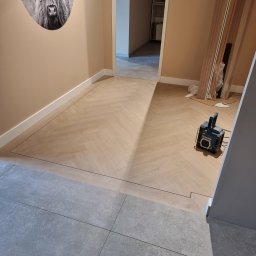 KONKRET Zbigniew Stańczak - Układanie parkietu w jodełkę przechodzące w szare płytki, widoczna lampa budowlana Makita i zdjęcie krowy na ścianie.