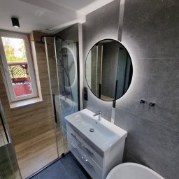 KONKRET Zbigniew Stańczak - Nowoczesna łazienka z szarą płytką imitującą kamień, okrągłym lustrem z podświetleniem LED, białą szafką z umywalką i czarną kabiną prysznicową z widokiem na balkon przez okno.