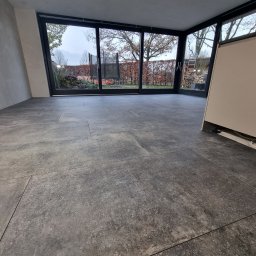 KONKRET Zbigniew Stańczak - Szare płytki podłogowe ułożone w salonie z dużym oknem tarasowym z widokiem na ogród w miejscowości Rudy, widoczna instalacja pod szafką.