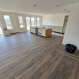 KONKRET Zbigniew Stańczak - Wnętrze w trakcie remontu: ułożona podłoga z paneli imitujących drewno, niedokończona zabudowa kuchenna, drabina przy oknie, widoczne narzędzia w wiadrze.