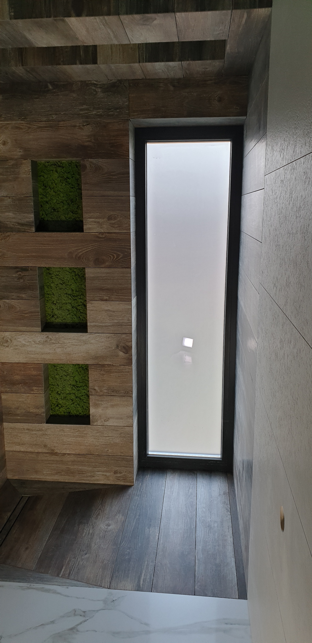 Wnętrze łazienki z drewnopodobnymi płytkami na ścianie i podłodze, pionowe okno z matową szybą oraz panele z mchem dekoracyjnym.