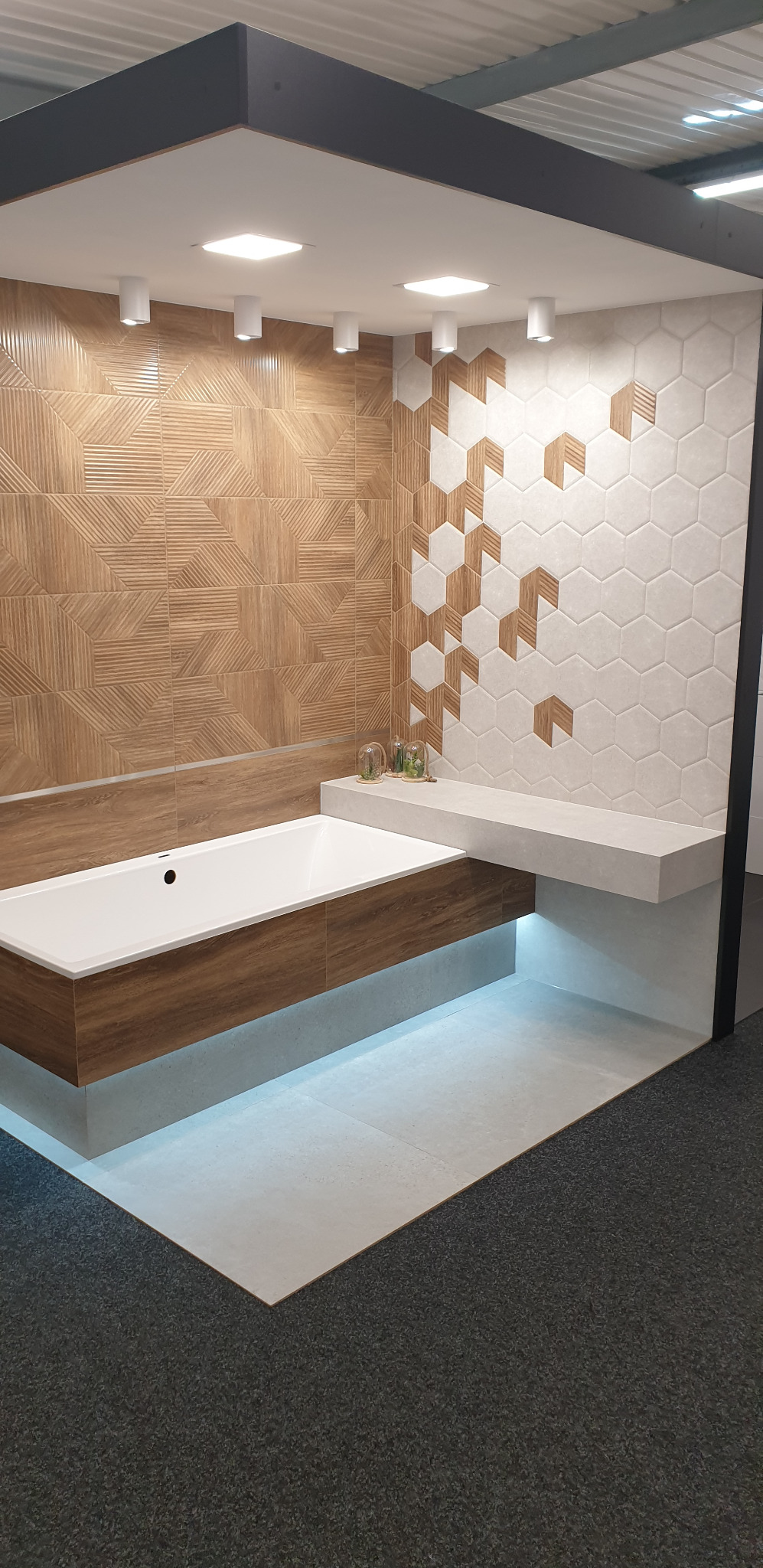 Nowoczesna aranżacja łazienki z wanną obudowaną drewnopodobnymi panelami, podświetleniem LED, geometrycznymi płytkami na ścianie i półką z dekoracjami.