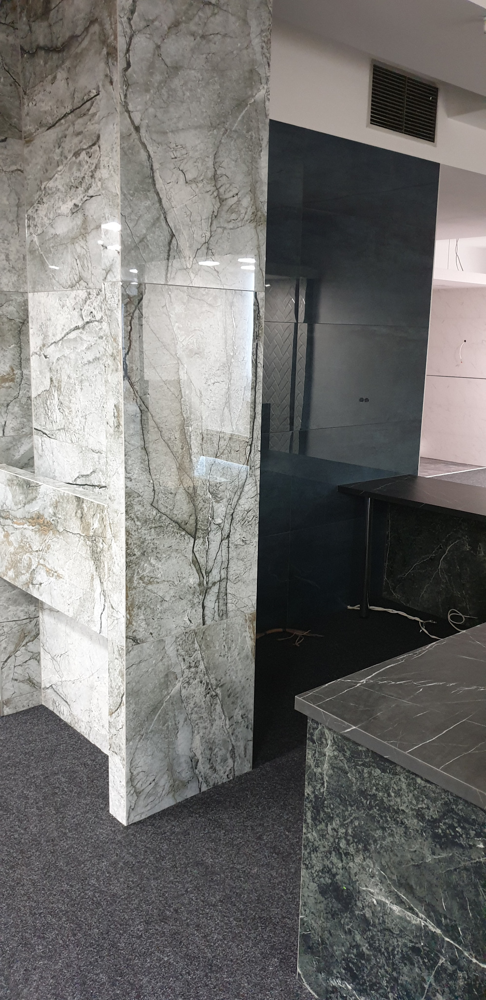 Wykończona ściana i fragment blatu w odcieniach szarości z marmurkowym wzorem, widoczna wentylacja sufitowa i fragment podłogi z ciemnej wykładziny.