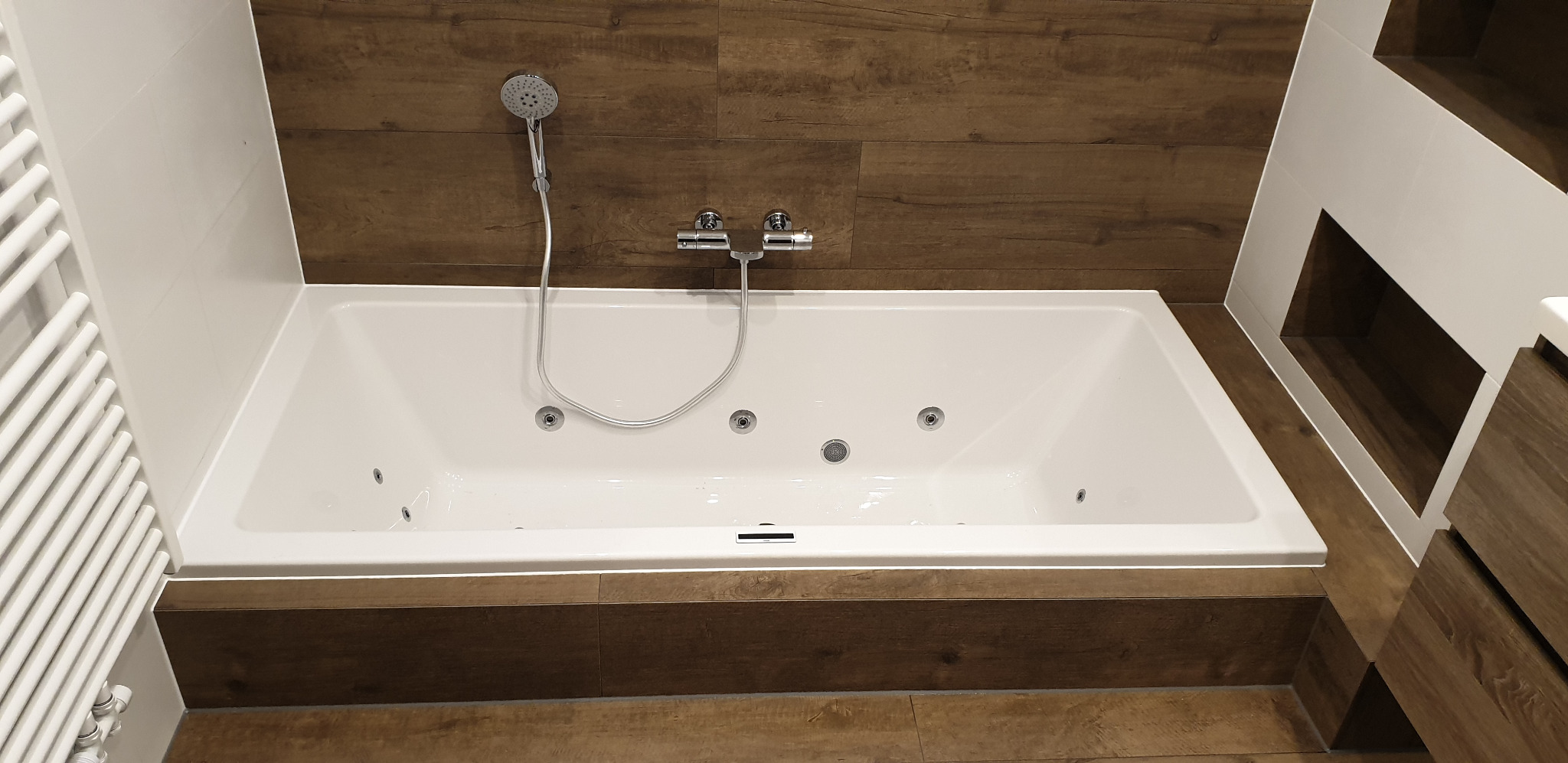 Biała wanna z hydromasażem, chromowana armatura, brązowe płytki imitujące drewno na ścianie i podłodze, biały grzejnik drabinkowy po lewej, nowoczesna aranżacja łazienki.