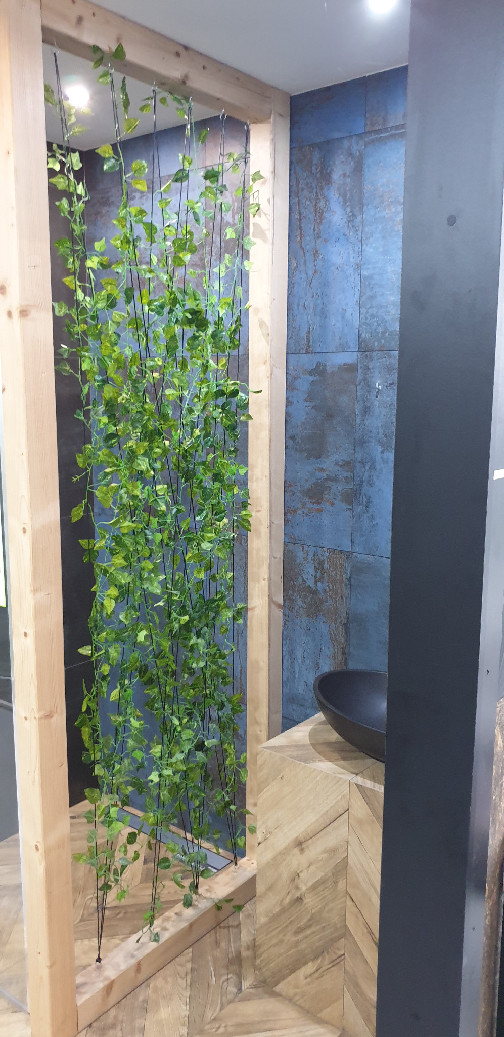 Aranżacja łazienki z drewnianą konstrukcją obłożoną sztuczną zielenią, umywalką nablatową i ścianą wyłożoną płytkami w odcieniach niebieskiego i brązu.