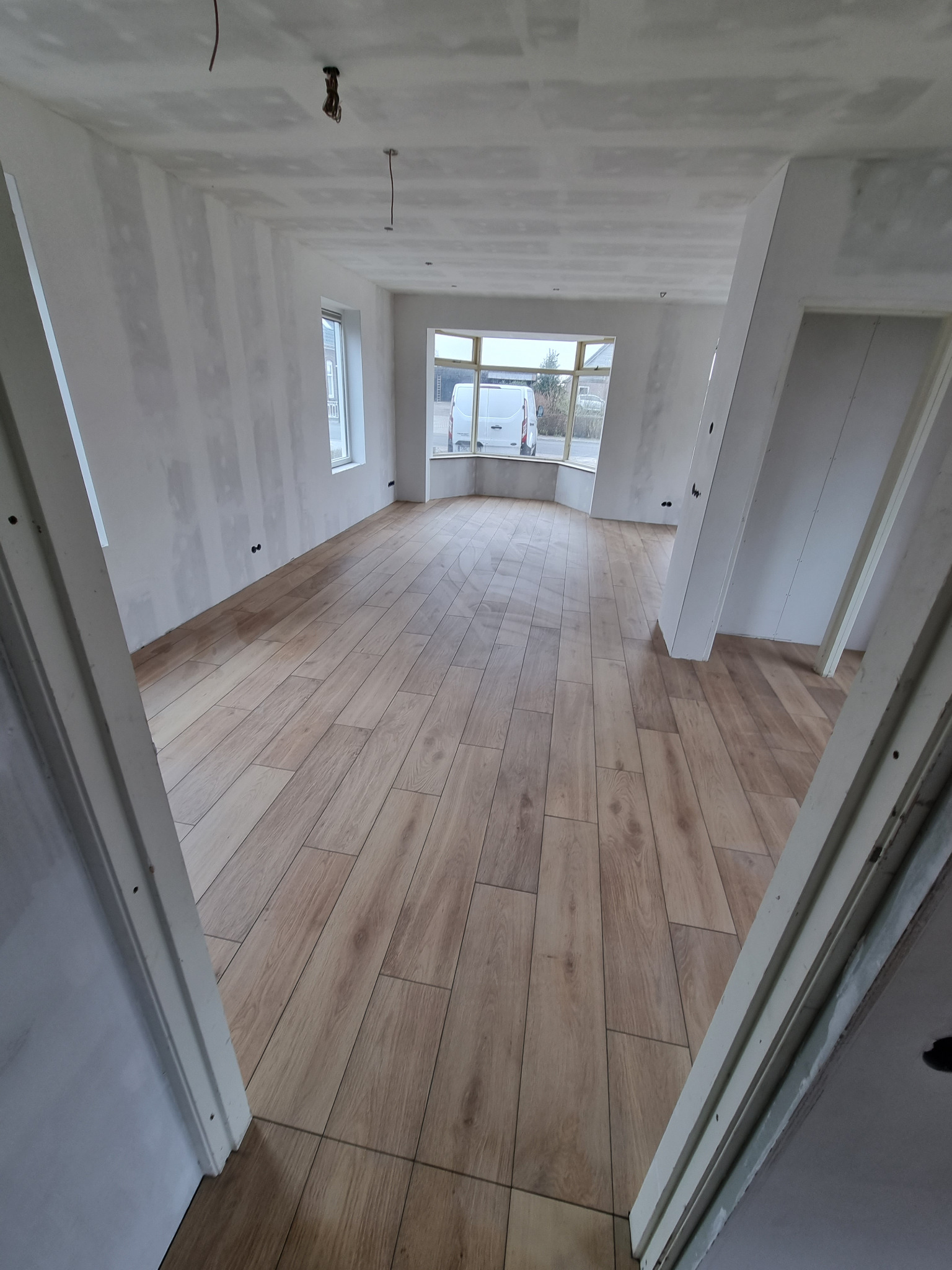Wnętrze pokoju w trakcie prac wykończeniowych: ściany po szpachlowaniu, ułożona podłoga z paneli imitujących drewno, widoczne kable zwisające z sufitu, okno z widokiem na ulicę.
