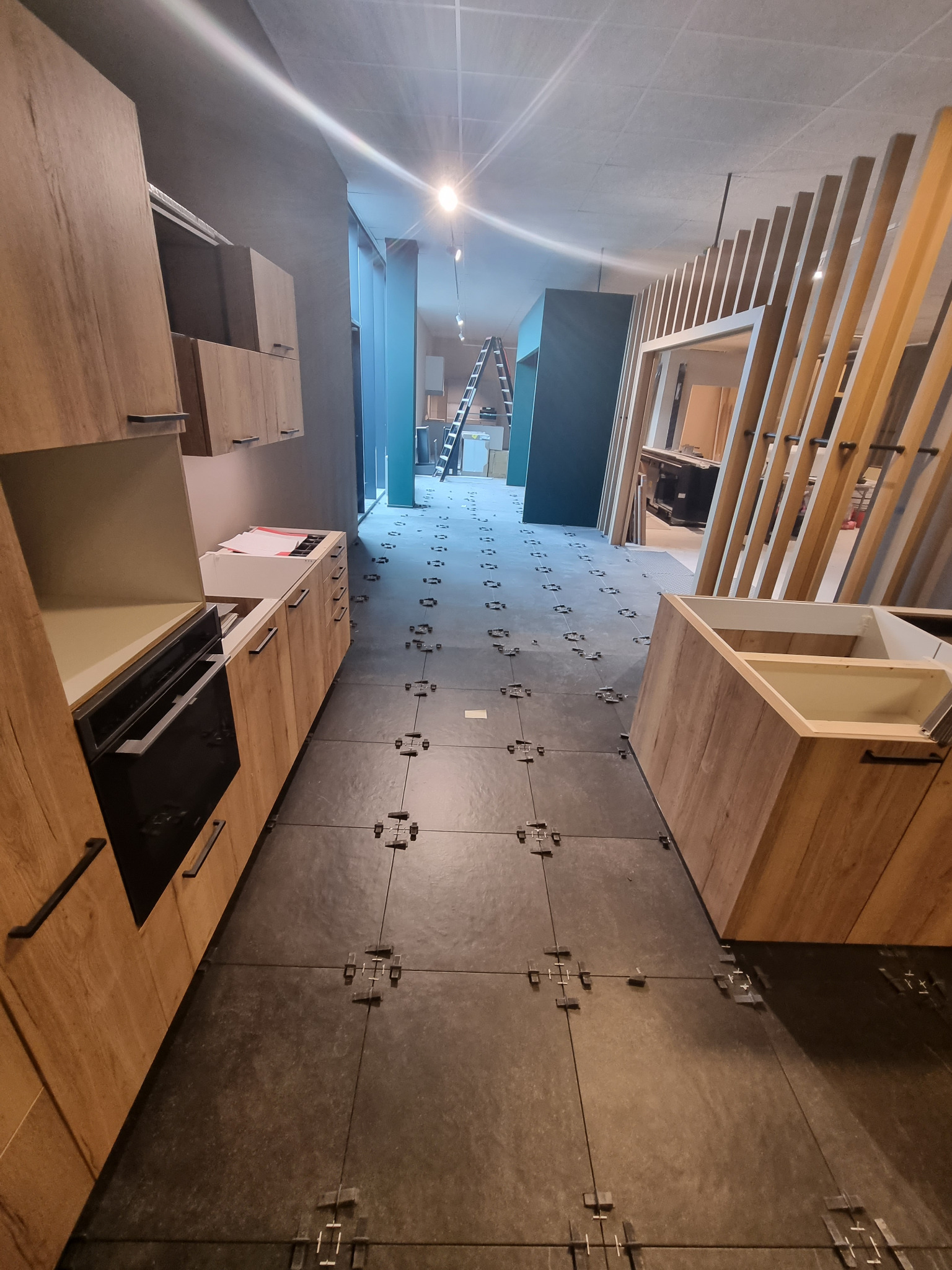 Montaż szarej podłogi z płytek w kuchni, widoczne klipsy poziomujące, częściowo zamontowane szafki z drewnopodobnym wykończeniem, drabina w tle.