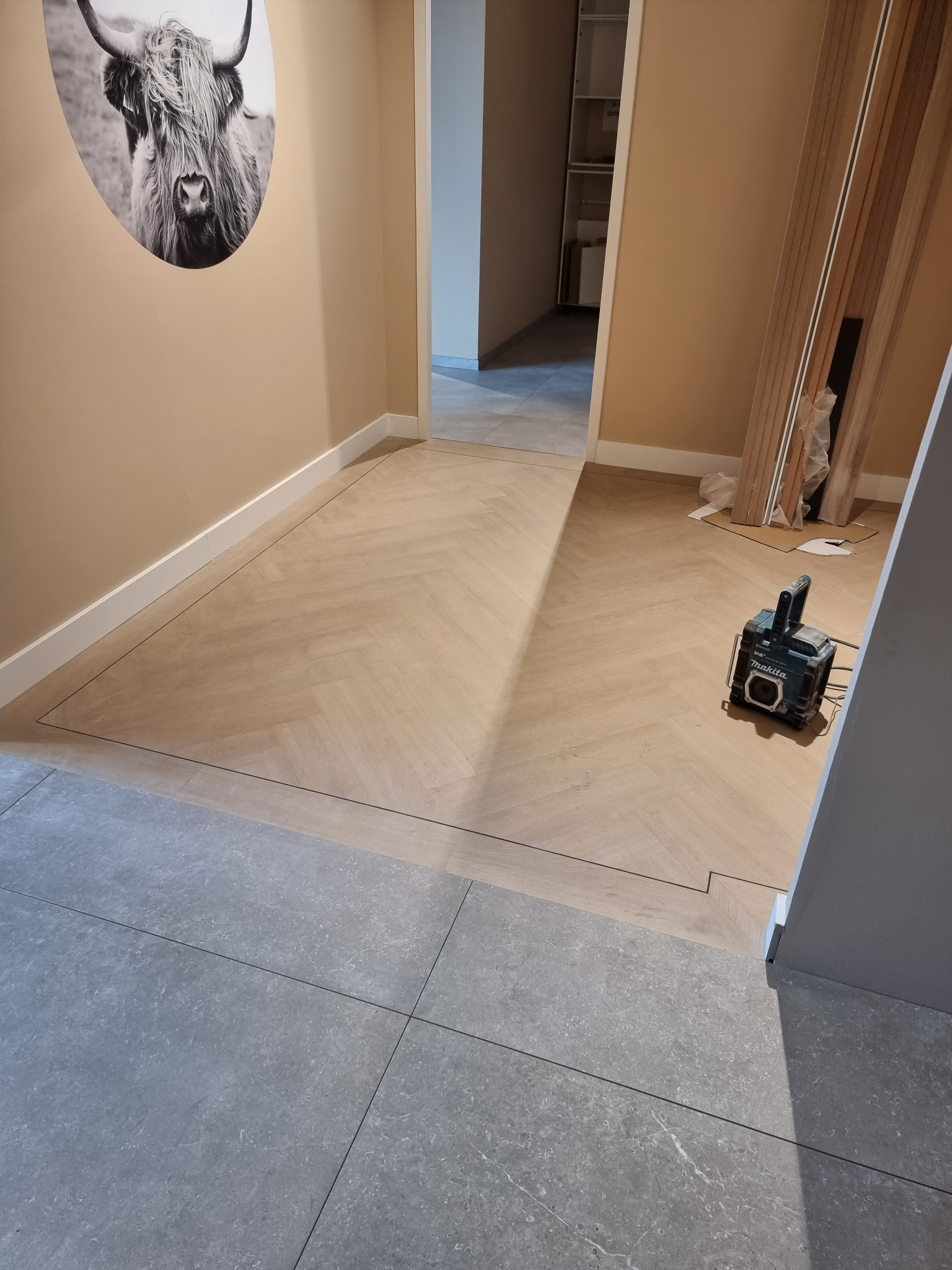 Układanie parkietu w jodełkę przechodzące w szare płytki, widoczna lampa budowlana Makita i zdjęcie krowy na ścianie.