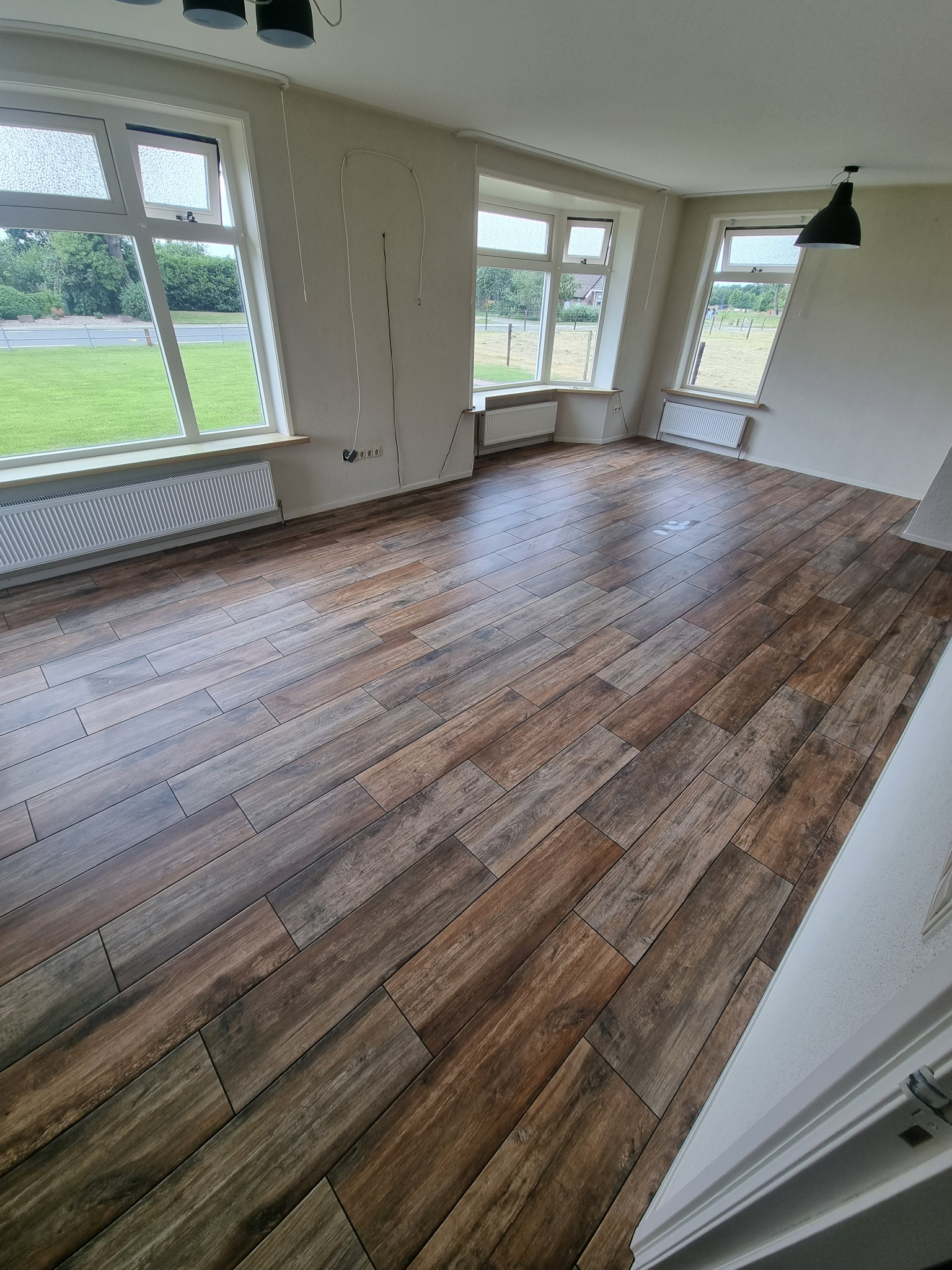 Ukończona podłoga z płytek imitujących drewno w jasnym, pustym pokoju z trzema oknami i białymi grzejnikami pod nimi.