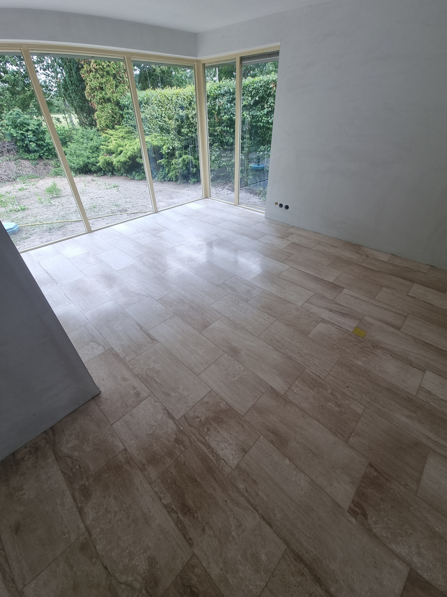 Ułożona podłoga z płytek imitujących trawertyn w jasnym pomieszczeniu z dużymi oknami wychodzącymi na zieleń.