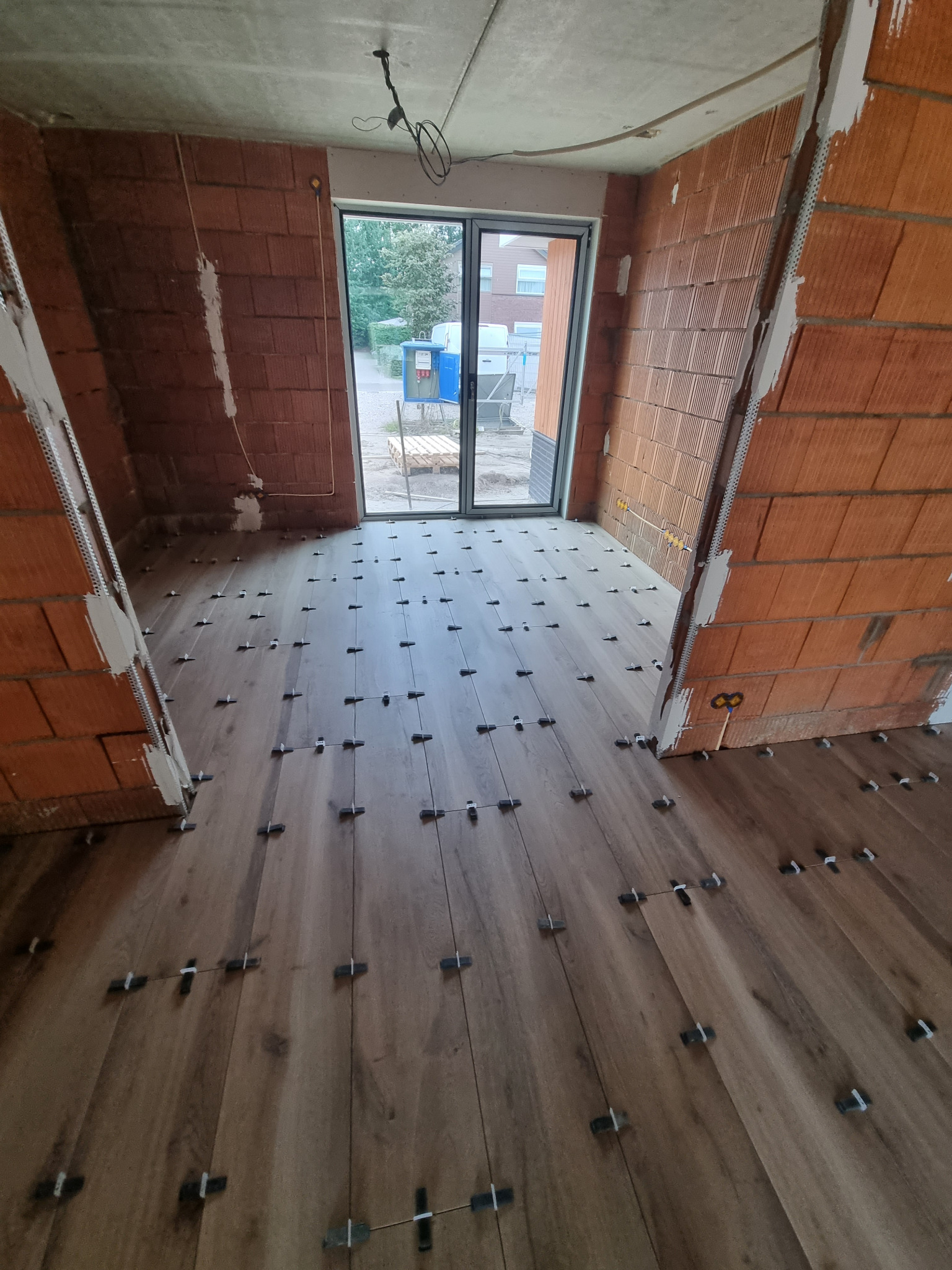 Układanie drewnopodobnych płytek podłogowych z użyciem krzyżyków dystansowych w surowym wnętrzu z ceglanymi ścianami i widokiem na zewnątrz przez przeszklone drzwi balkonowe.