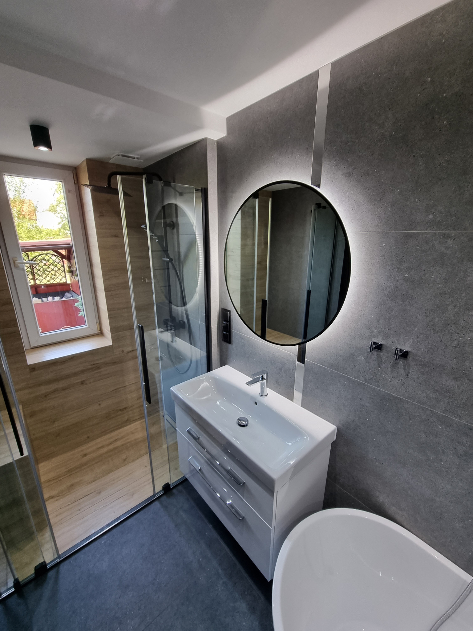 Nowoczesna łazienka z szarą płytką imitującą kamień, okrągłym lustrem z podświetleniem LED, białą szafką z umywalką i czarną kabiną prysznicową z widokiem na balkon przez okno.