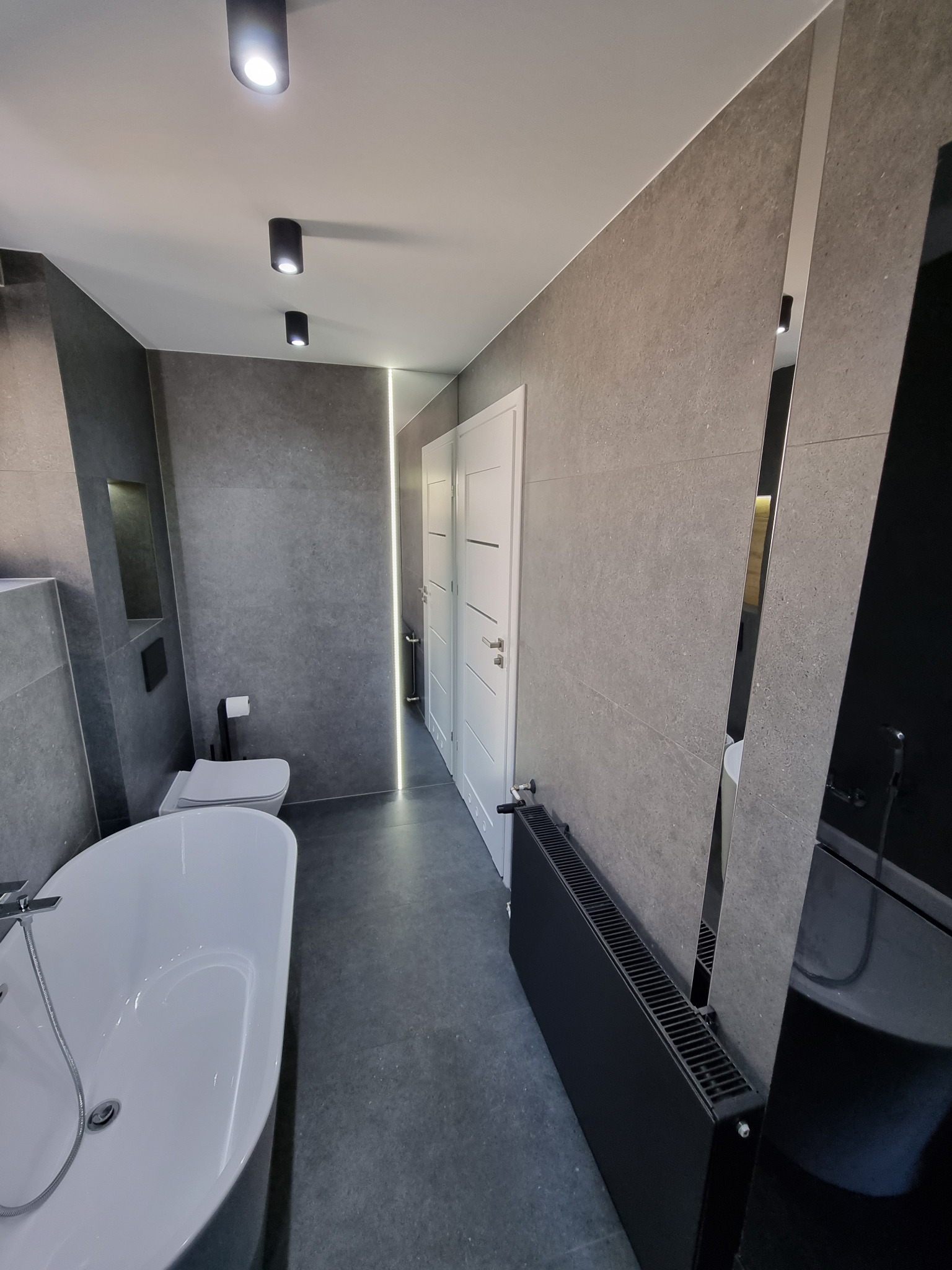 Nowoczesna łazienka w odcieniach szarości z wolnostojącą białą wanną, podwieszaną toaletą, czarnym grzejnikiem i pionowym oświetleniem LED w szczelinie między ścianą a drzwiami.