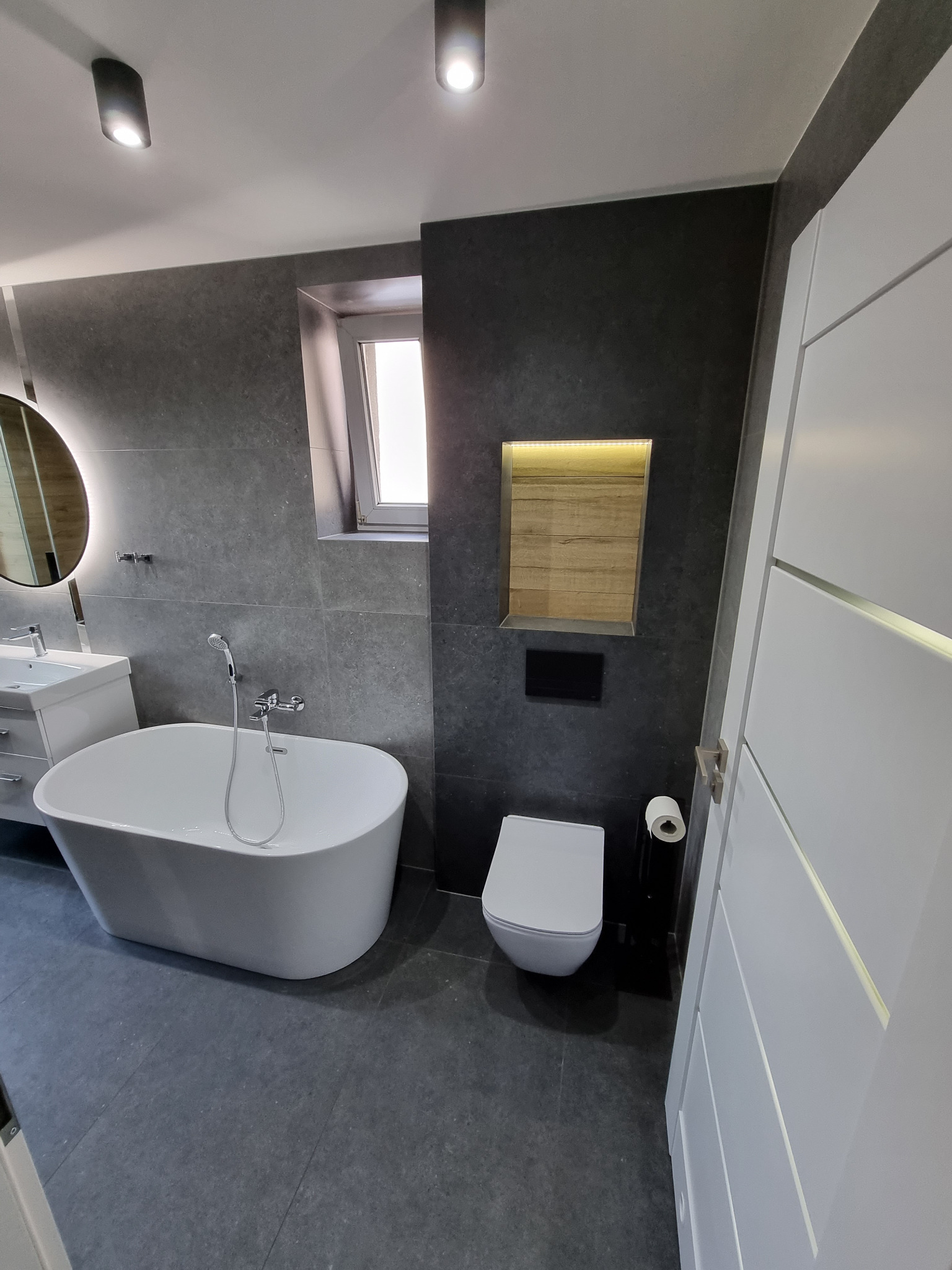 Nowoczesna łazienka z wolnostojącą wanną, szarą płytką imitującą kamień, podświetlaną niszą z drewnianym akcentem i białymi drzwiami z frezowanymi panelami.