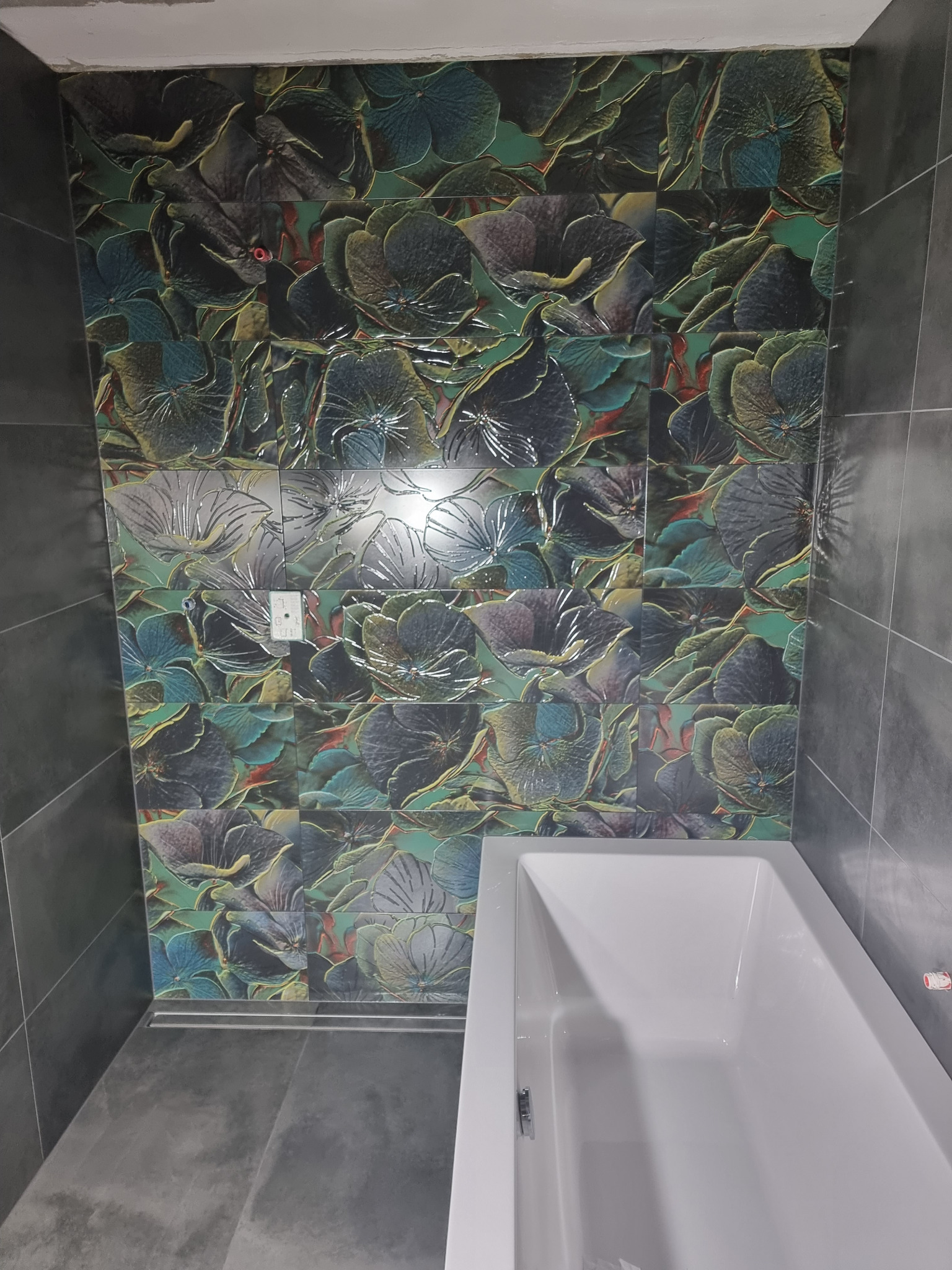 Wanna w łazience wyłożonej ciemnoszarą płytką i dekoracyjną mozaiką z motywem kwiatowym na ścianie, widoczny fragment gniazdka elektrycznego.