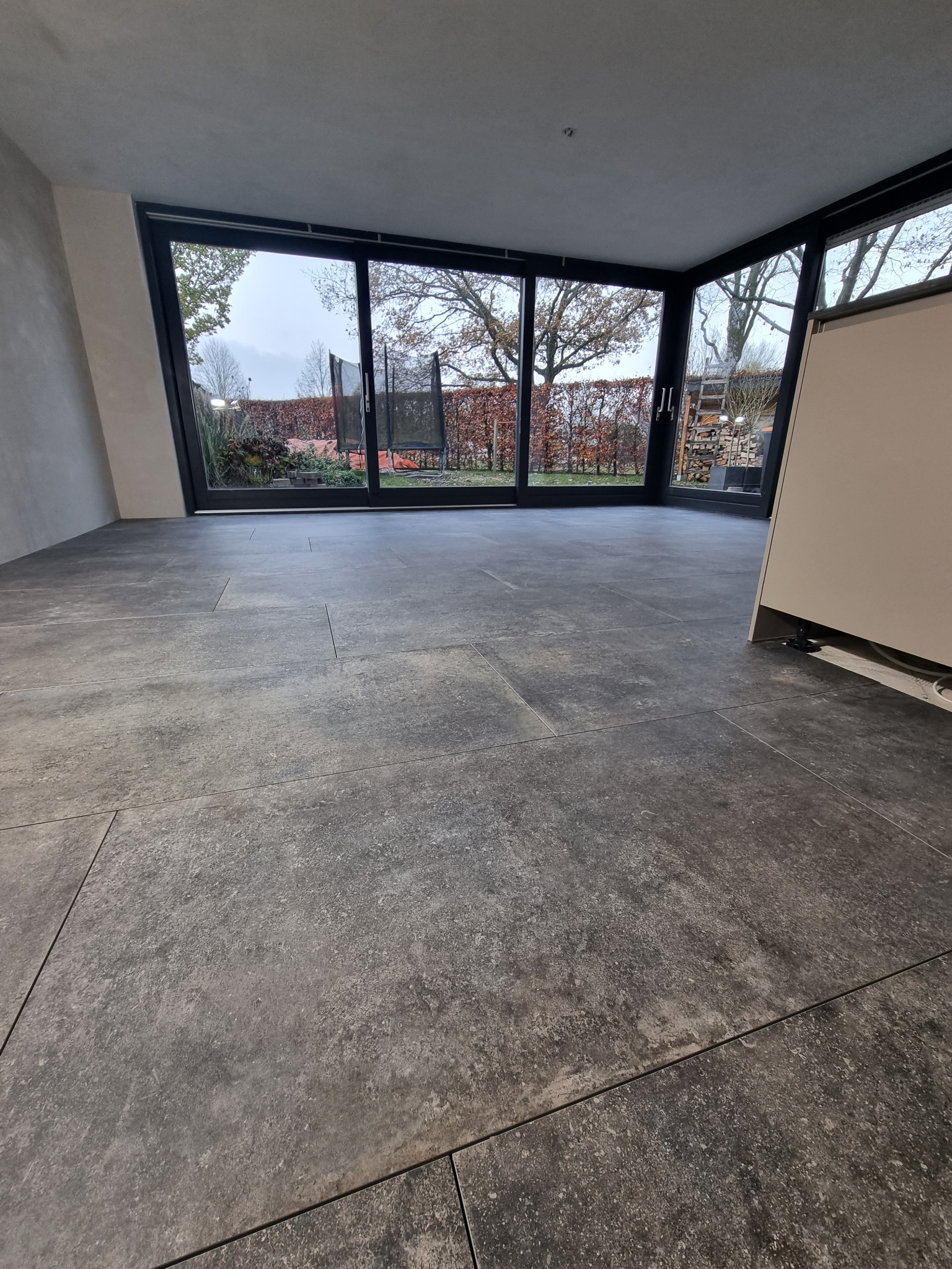 Szare płytki podłogowe ułożone w salonie z dużym oknem tarasowym z widokiem na ogród w miejscowości Rudy, widoczna instalacja pod szafką.