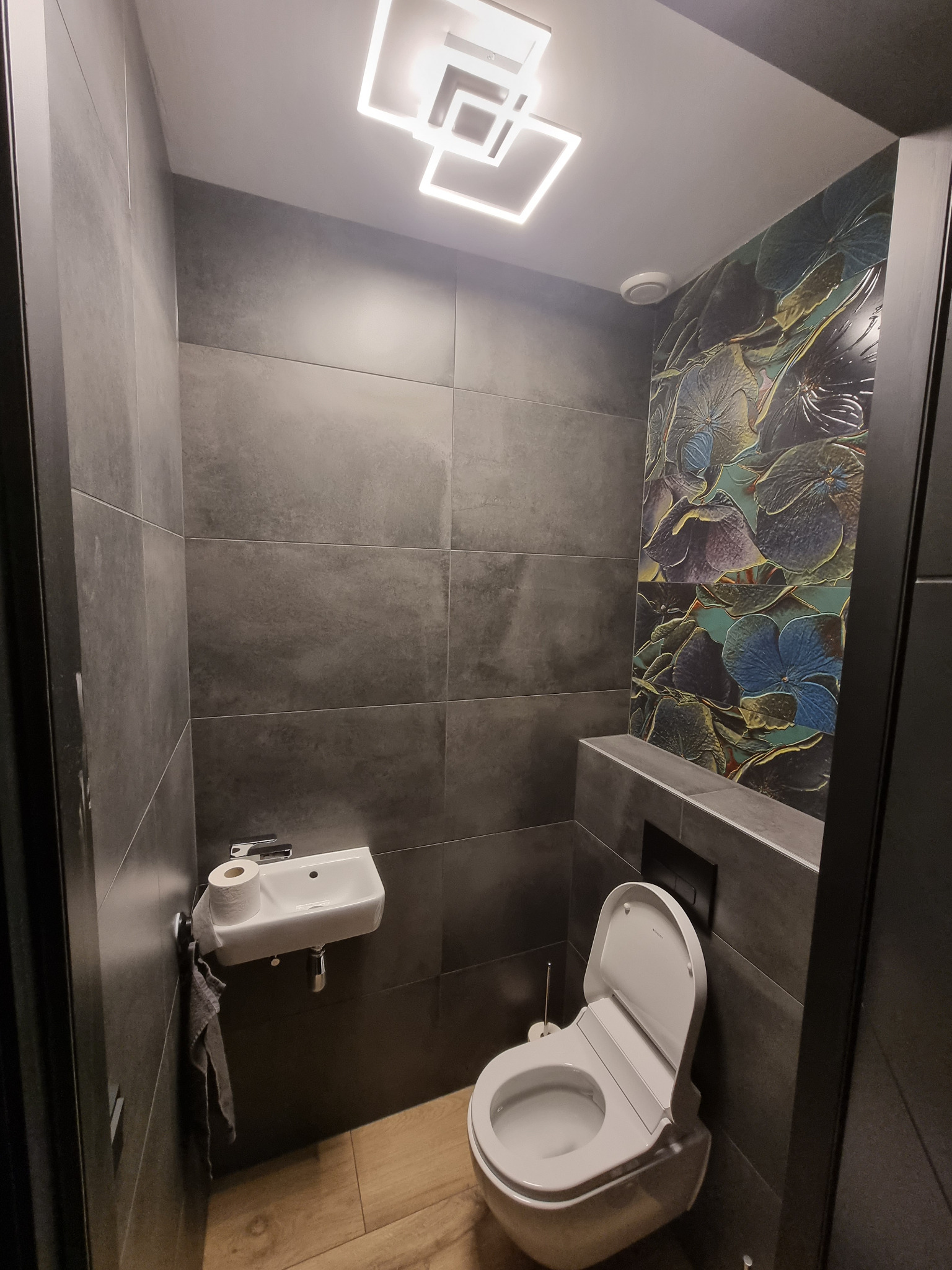 Nowoczesna, mała łazienka z szarymi płytkami wielkoformatowymi, podwieszaną toaletą z deską myjącą, małą umywalką narożną z szarą ściereczką i dekoracyjną ścianą z motywem kwiatowym.