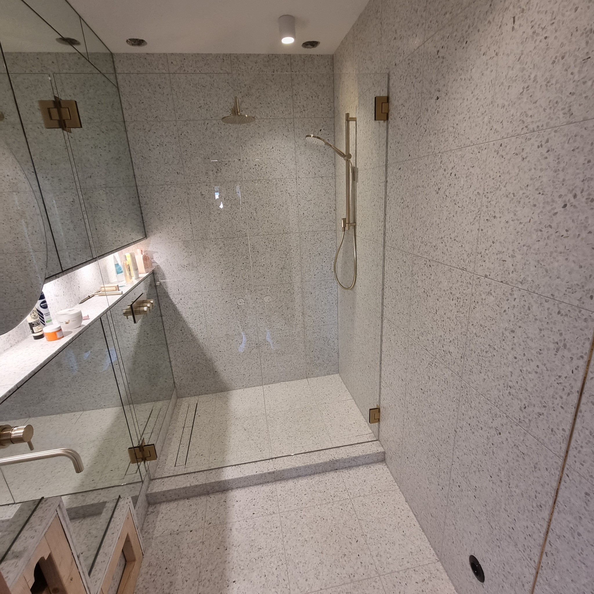 Nowoczesna kabina prysznicowa z szarymi płytkami lastryko, złotymi detalami armatury i szklanymi drzwiami, widok na półkę z kosmetykami wbudowaną w lustro.