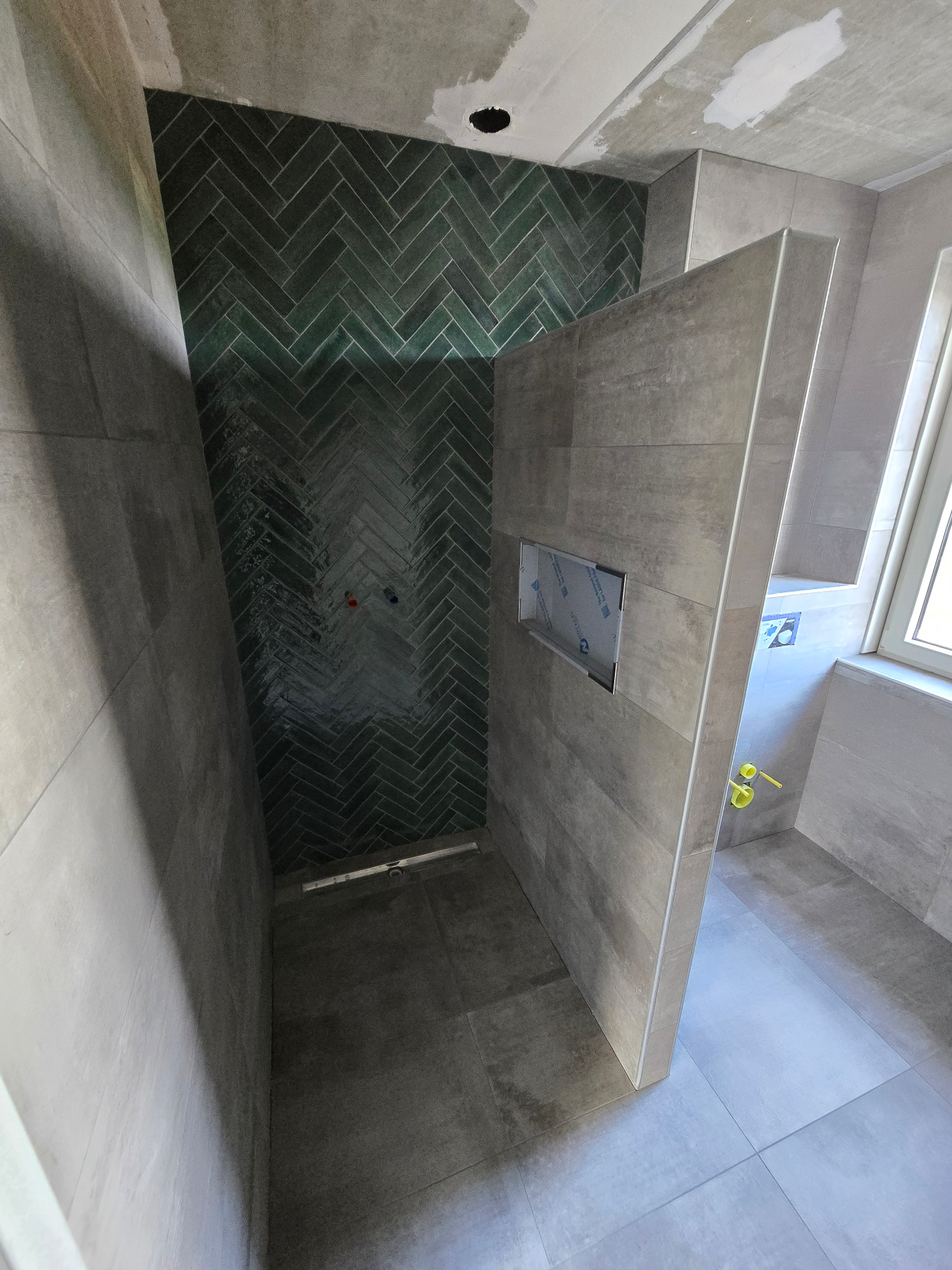 Niedokończona kabina prysznicowa z zielonymi płytkami ułożonymi w jodełkę na ścianie, szare płytki na podłodze i ścianach, widoczne rury i otwór na armaturę.