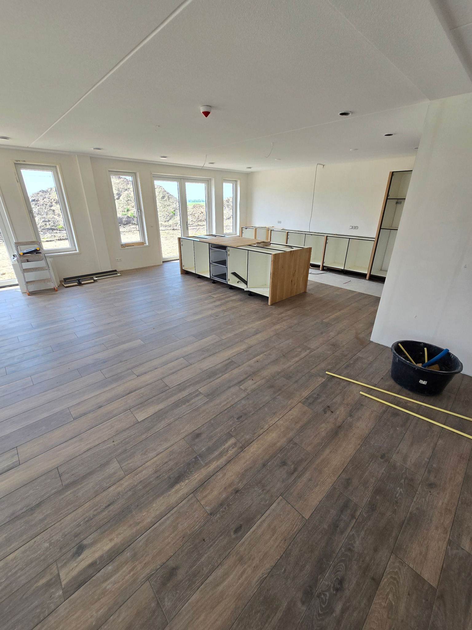 Wnętrze w trakcie remontu: ułożona podłoga z paneli imitujących drewno, niedokończona zabudowa kuchenna, drabina przy oknie, widoczne narzędzia w wiadrze.