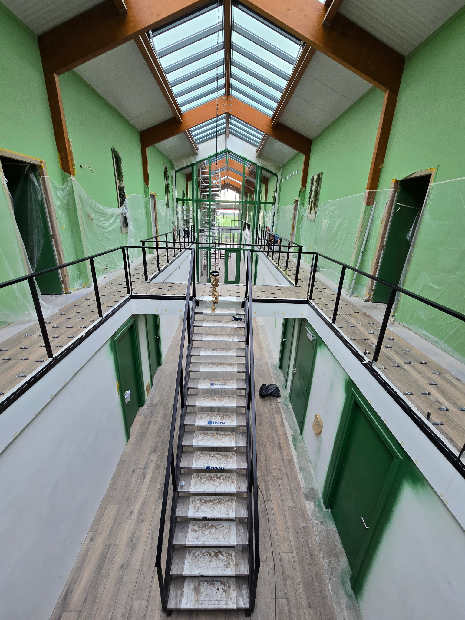 Wnętrze budynku w trakcie prac wykończeniowych: świeżo pomalowane na zielono ściany, drewniana konstrukcja dachu z przeszkleniami, schody z zabezpieczeniami na stopniach, podłoga wyłożona panelami,...