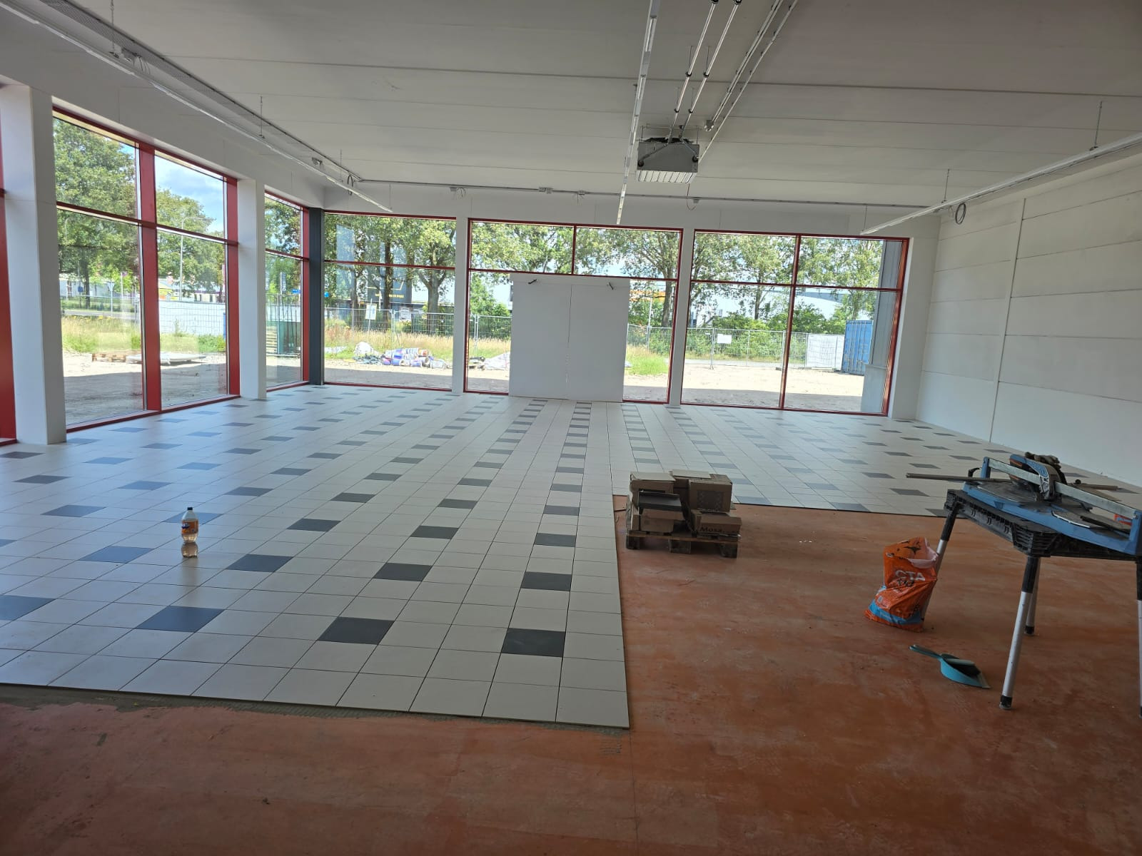 Układanie jasnych płytek podłogowych z ciemnymi akcentami w przestronnym, jasnym pomieszczeniu z dużymi oknami; widoczny fragment nieprzygotowanej podłogi, paleta z płytkami i przecinarka do płytek.