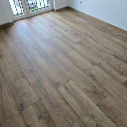 REMONTY PK TOMASZEWSCY SPÓŁKA Z OGRANICZONĄ ODPOWIEDZIALNOŚCIĄ - Ukończona podłoga z paneli o wzorze drewna, ułożona w jasnym pokoju z białymi ścianami i grzejnikiem przy oknie balkonowym, widok pod kątem.