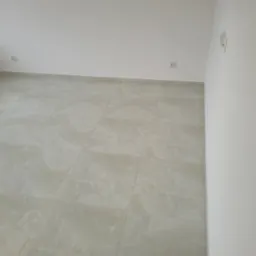 Wykończone wnętrze: jasne płytki podłogowe z subtelnym wzorem marmuru, gładkie białe ściany z zamontowanymi gniazdkami i włącznikami światła. Minimalistyczny design.