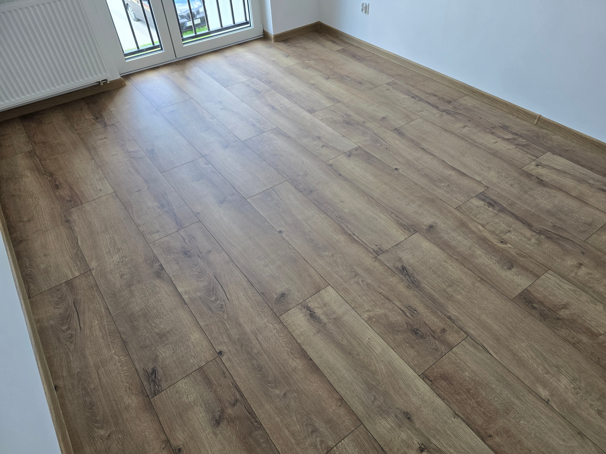 Ukończona podłoga z paneli o wzorze drewna, ułożona w jasnym pokoju z białymi ścianami i grzejnikiem przy oknie balkonowym, widok pod kątem.