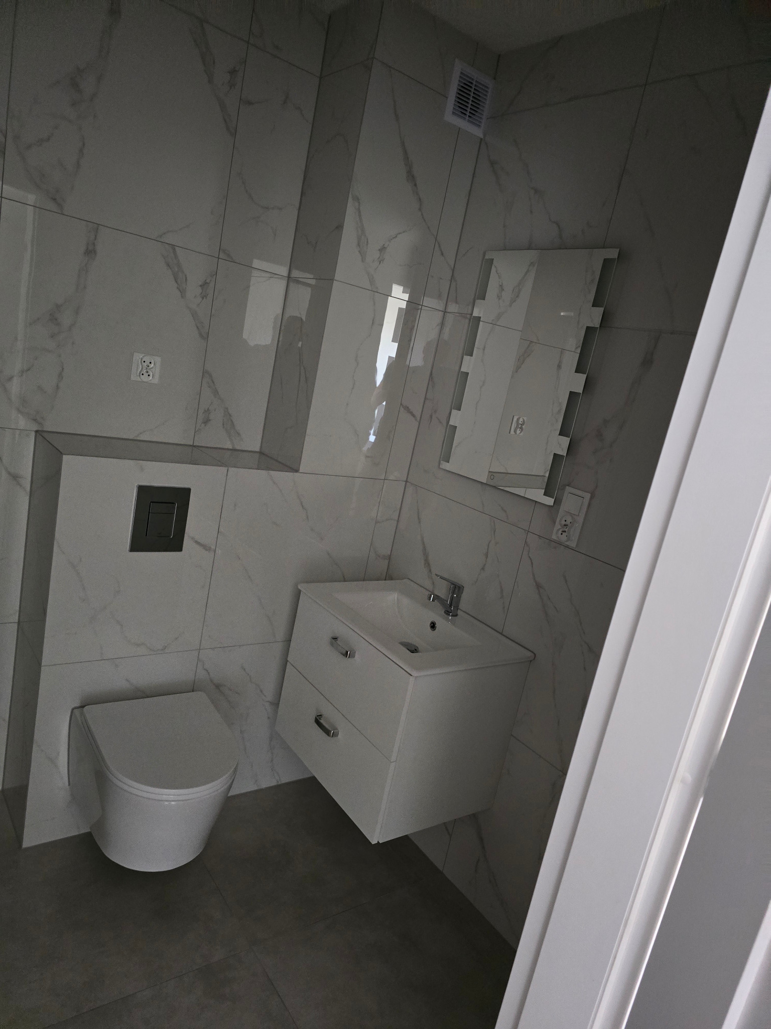 Nowoczesna łazienka z marmurowymi płytkami, wiszącą toaletą i białą szafką umywalkową. Minimalistyczny design, szara podłoga i lustro z oświetleniem.