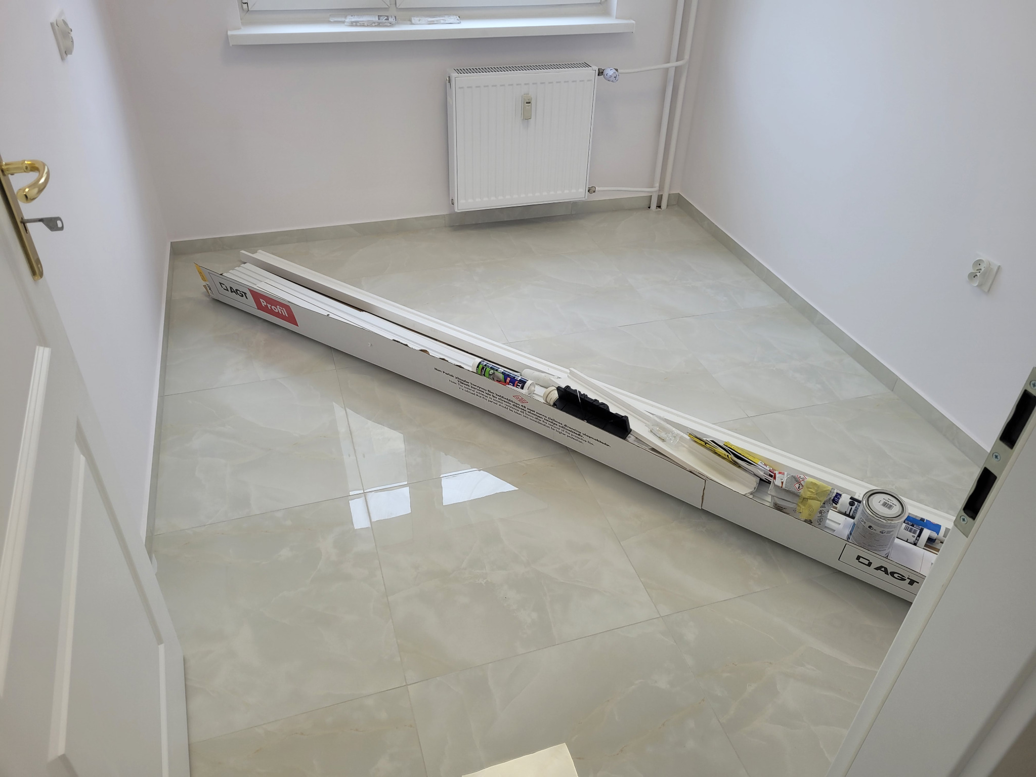 Jasne wnętrze z błyszczącą podłogą. W centralnej części karton z listwami przypodłogowymi AGT Profil i akcesoriami do montażu. Widoczny fragment okna i grzejnika.