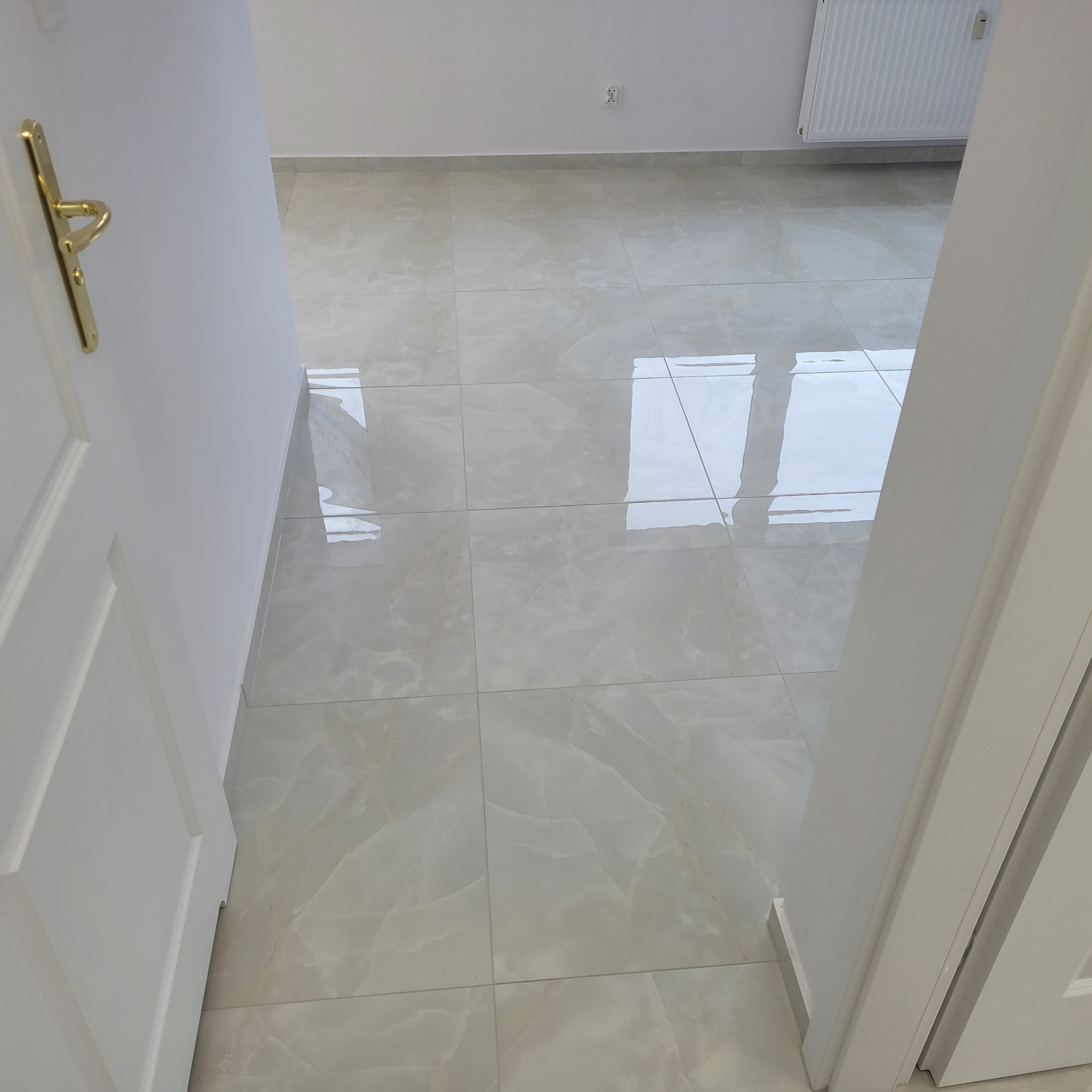 Widok przez otwarte drzwi na błyszczące, jasne płytki podłogowe w pokoju z białymi ścianami i grzejnikiem. Złota klamka na białych drzwiach.