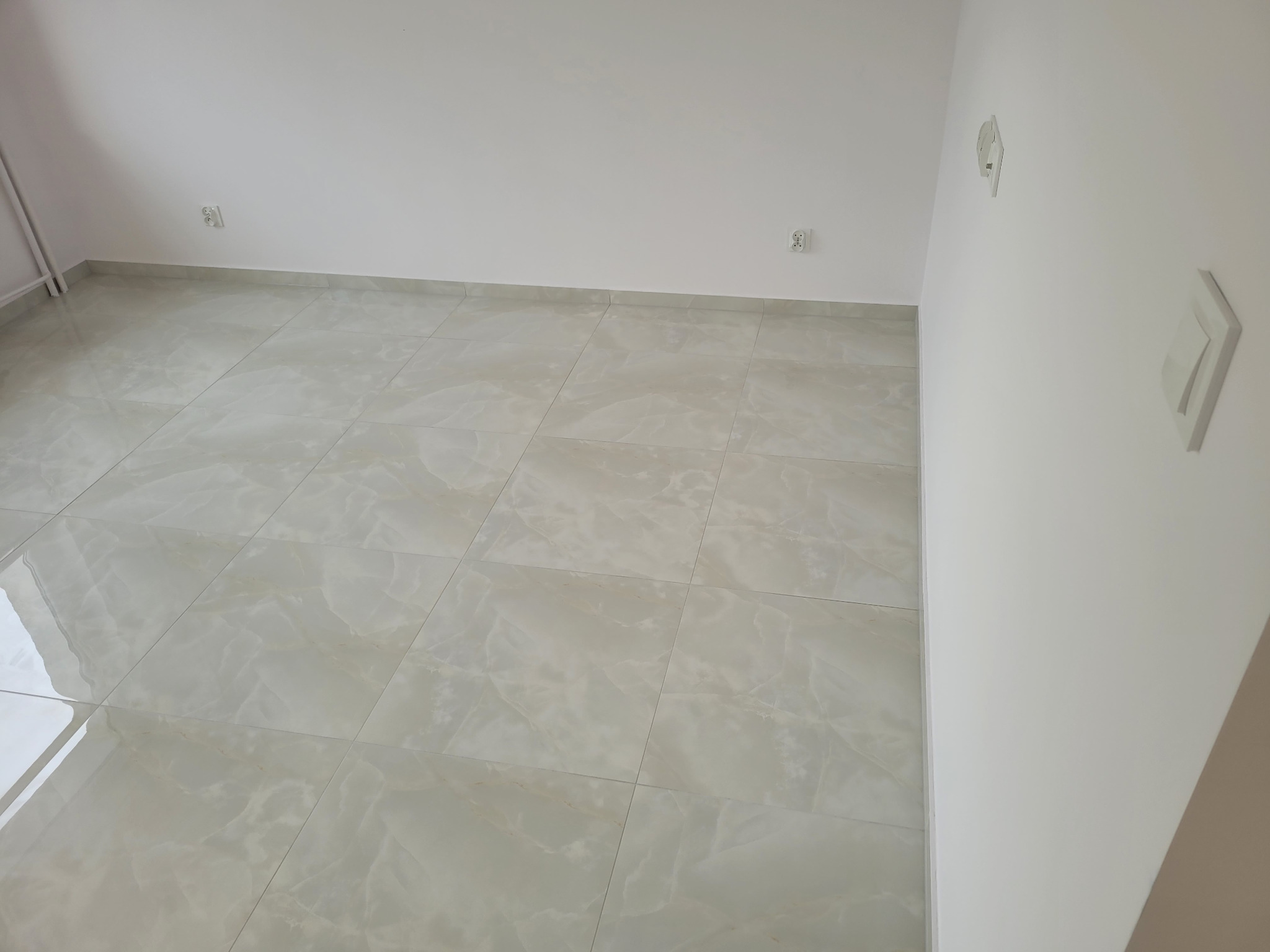 Wykończone wnętrze: jasne płytki podłogowe z subtelnym wzorem marmuru, gładkie białe ściany z zamontowanymi gniazdkami i włącznikami światła. Minimalistyczny design.