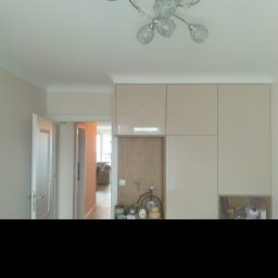 JARDAR SPÓŁKA Z OGRANICZONĄ ODPOWIEDZIALNOŚCIĄ - Nowoczesne wnętrze: błyszczące, beżowe szafy w zabudowie pod sufit, dekoracyjna lampa sufitowa z ażurowymi kloszami, otwarte drzwi do pokoju z kanapą w tle.