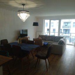 JARDAR SPÓŁKA Z OGRANICZONĄ ODPOWIEDZIALNOŚCIĄ - Jasne, przestronne wnętrze mieszkania po remoncie: parkiet, gładkie ściany, kryształowy żyrandol nad stołem z niebieskim obrusem, sofa z widokiem na balkon.